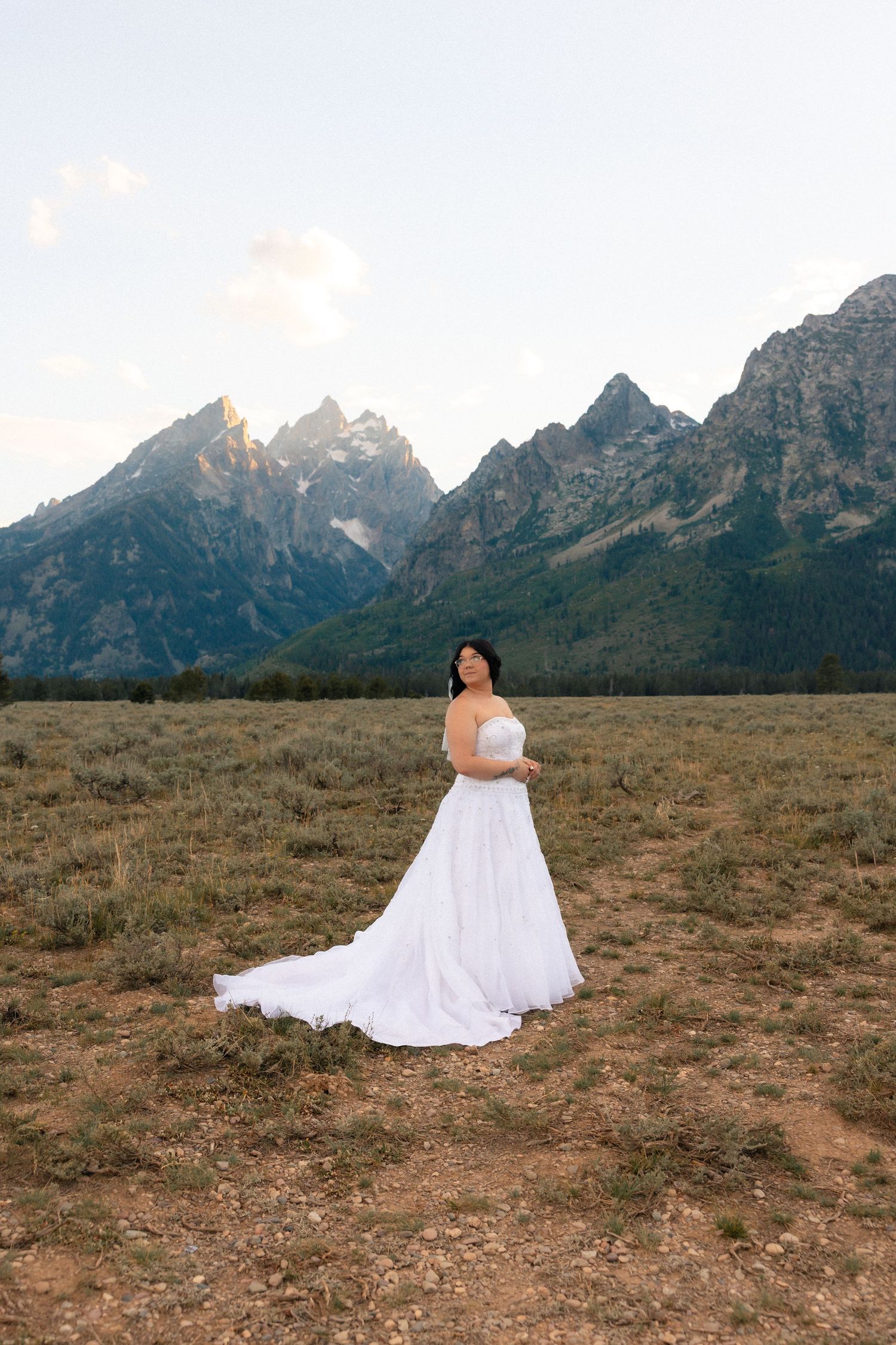 Bride in white gown poses in mountain meadow with Grand Teton peaks backdrop at dusk.