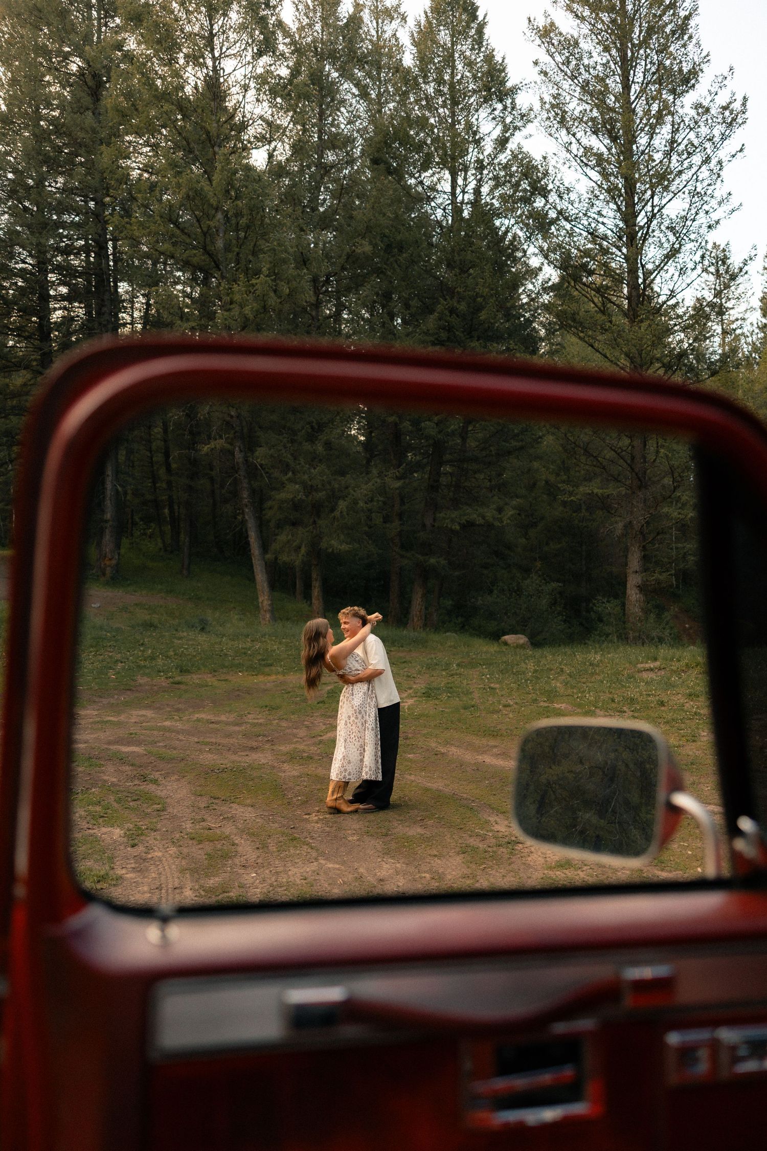 Couple embracing in a forest clearing, viewed through the window of a vintage red truck.