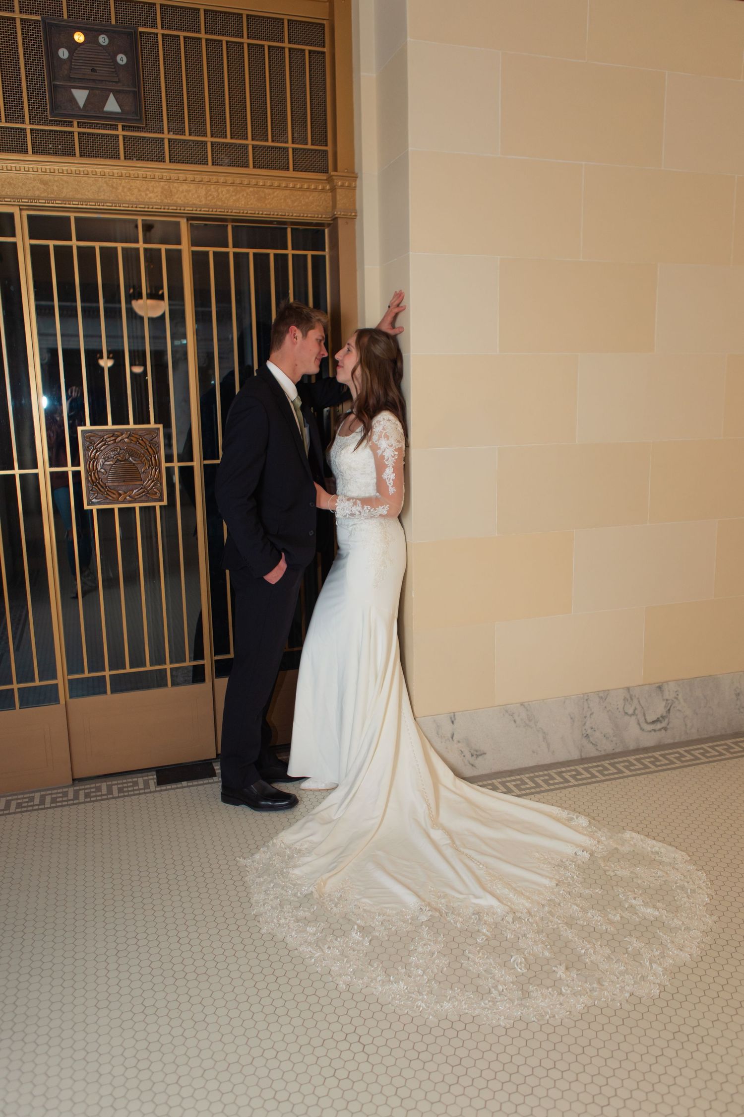 Bride in lace gown with cathedral train leans against wall with groom in tuxedo in elegant marble building.