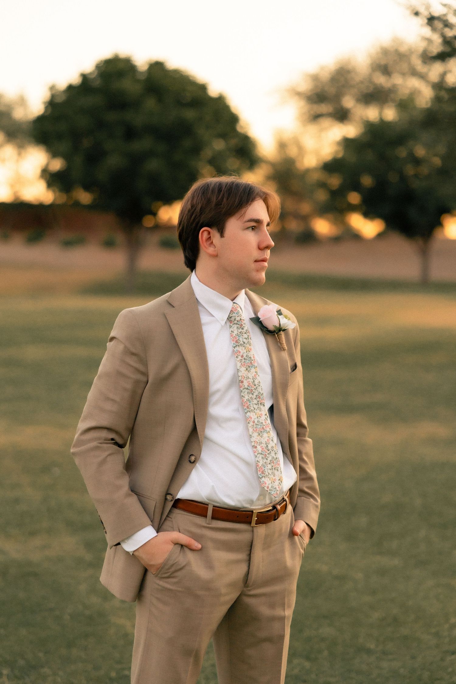 Young groom in tan suit with floral tie and boutonniere standing in golden hour sunlight at outdoor wedding venue.