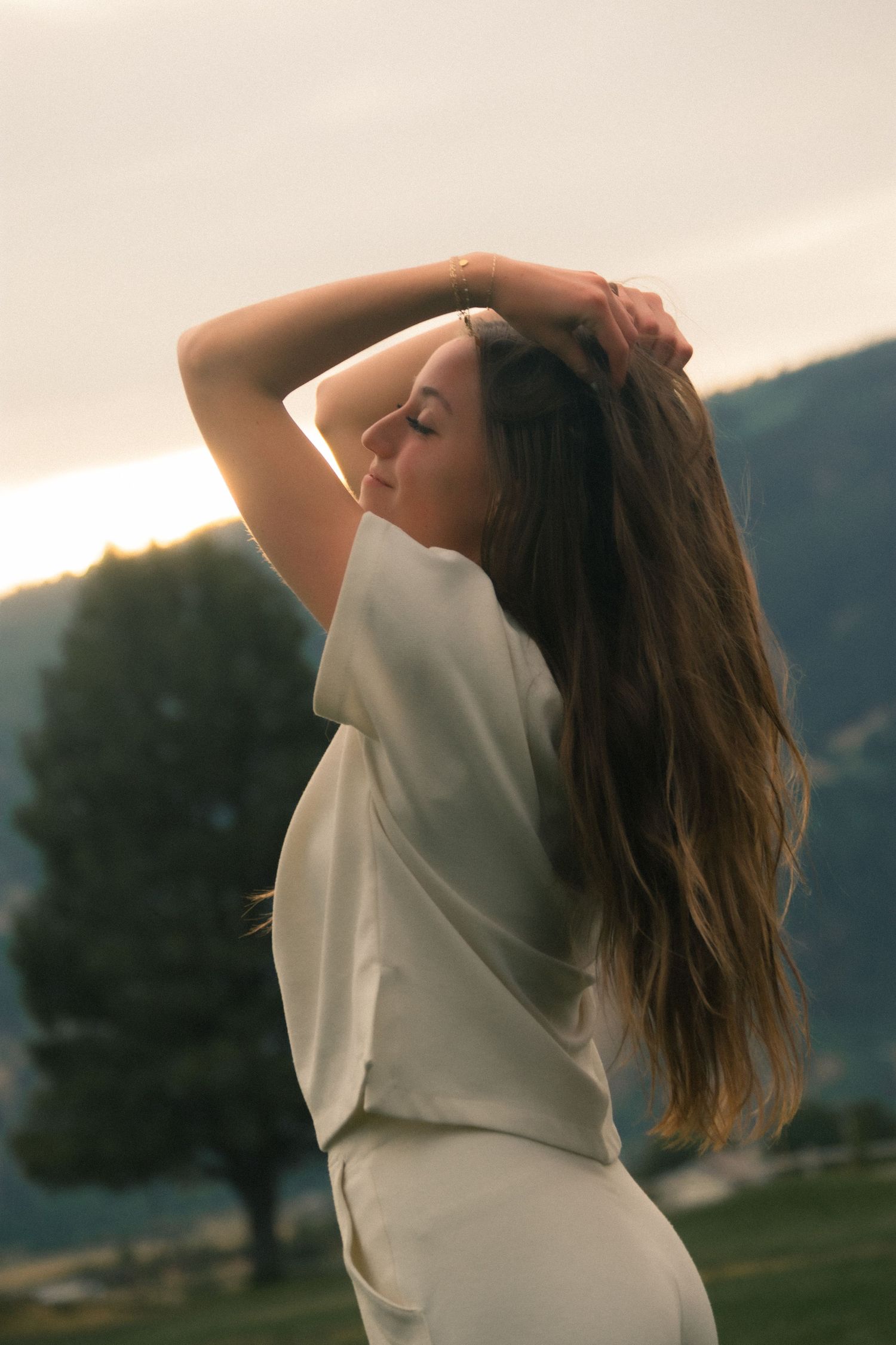 Young woman in white outfit holds her long hair up, posing outdoors at sunset with mountains in background.