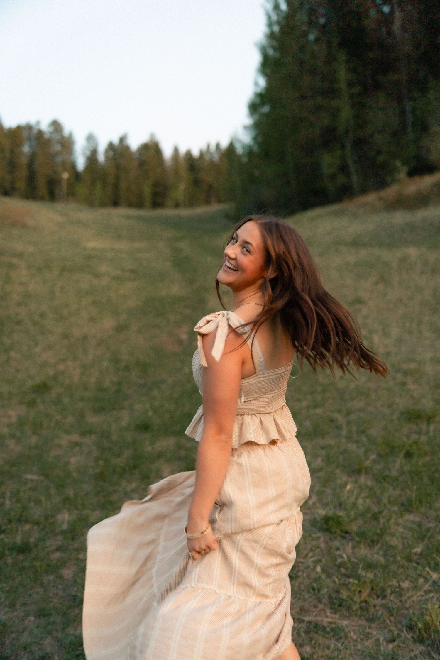 Smiling young woman in a flowing beige dress twirling in a green meadow with tall pine trees in the background.