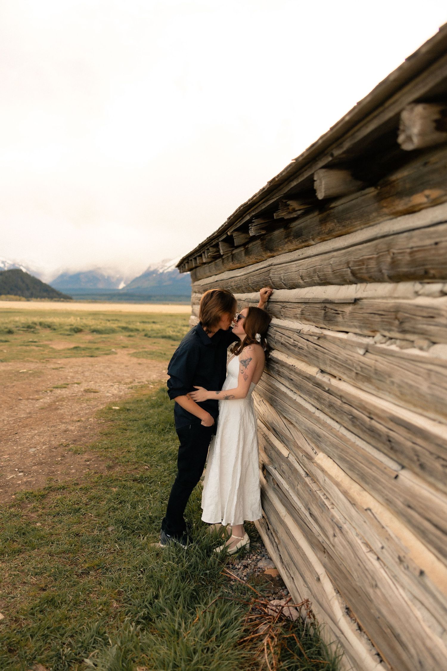 Couple sharing a romantic kiss against a rustic wooden barn wall with mountain landscape backdrop.