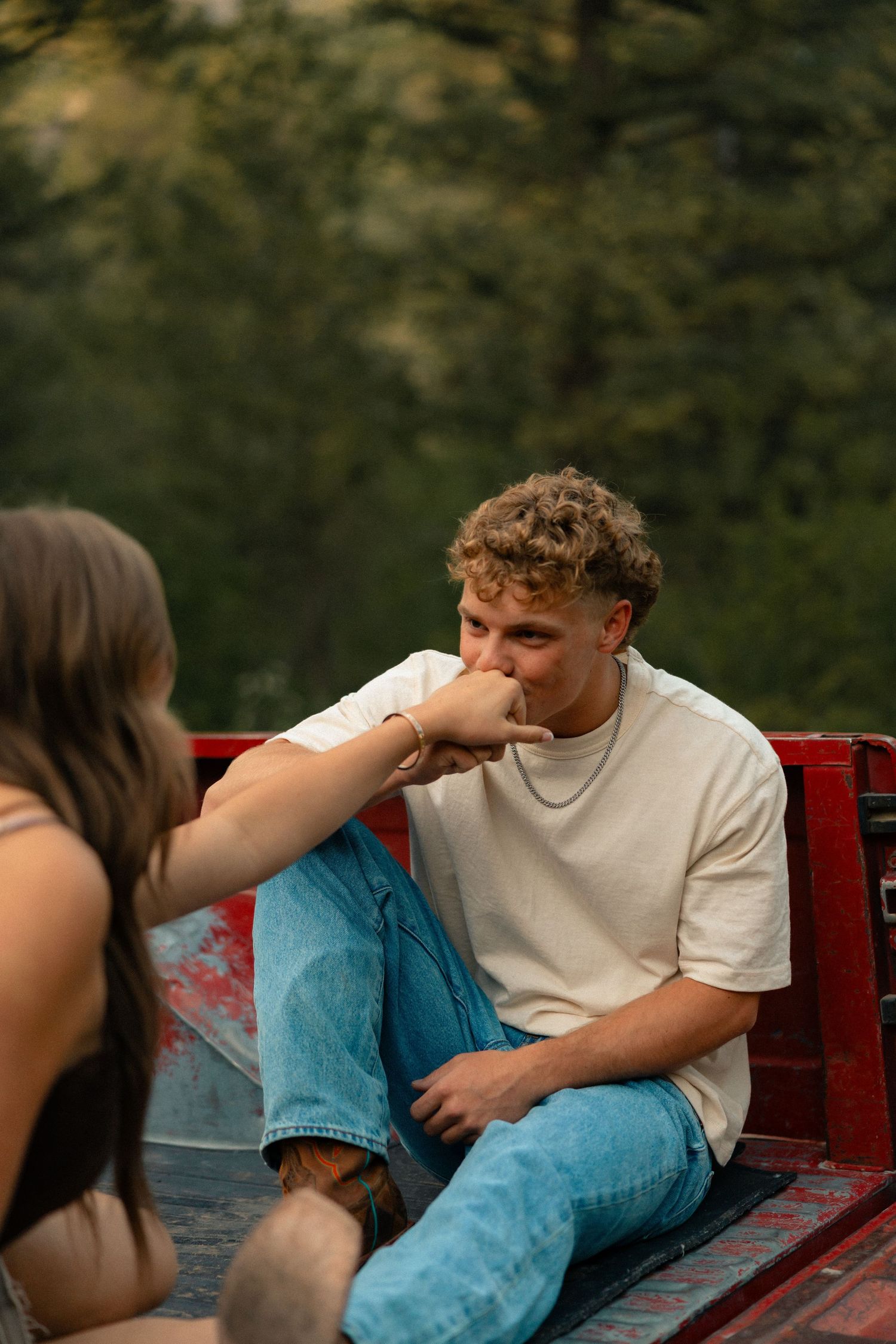 Young man kissing a woman's hand while sitting on a red truck bed with green trees in the background.