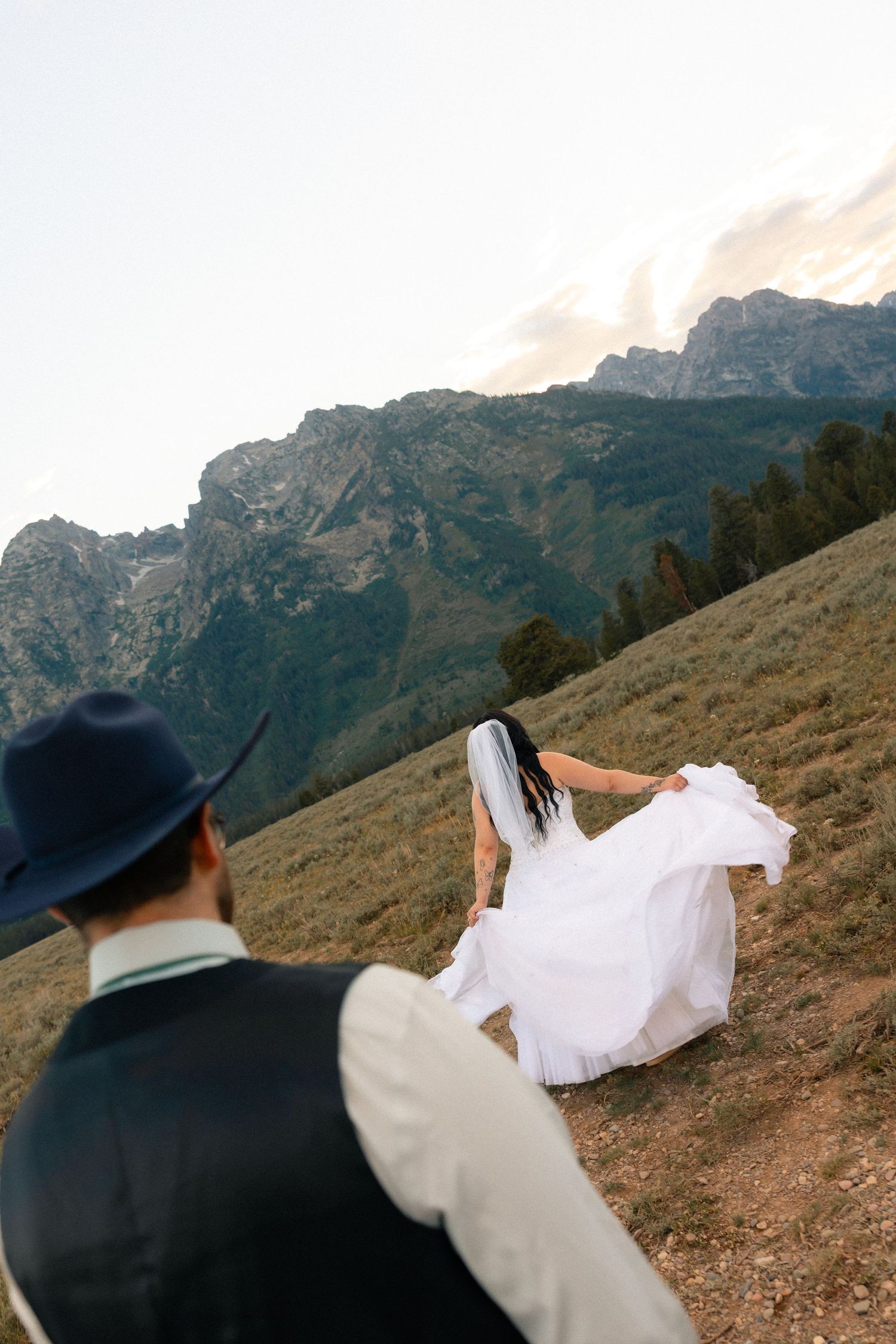 Bride in white gown and veil walks toward mountains as groom in navy hat follows during outdoor wedding.