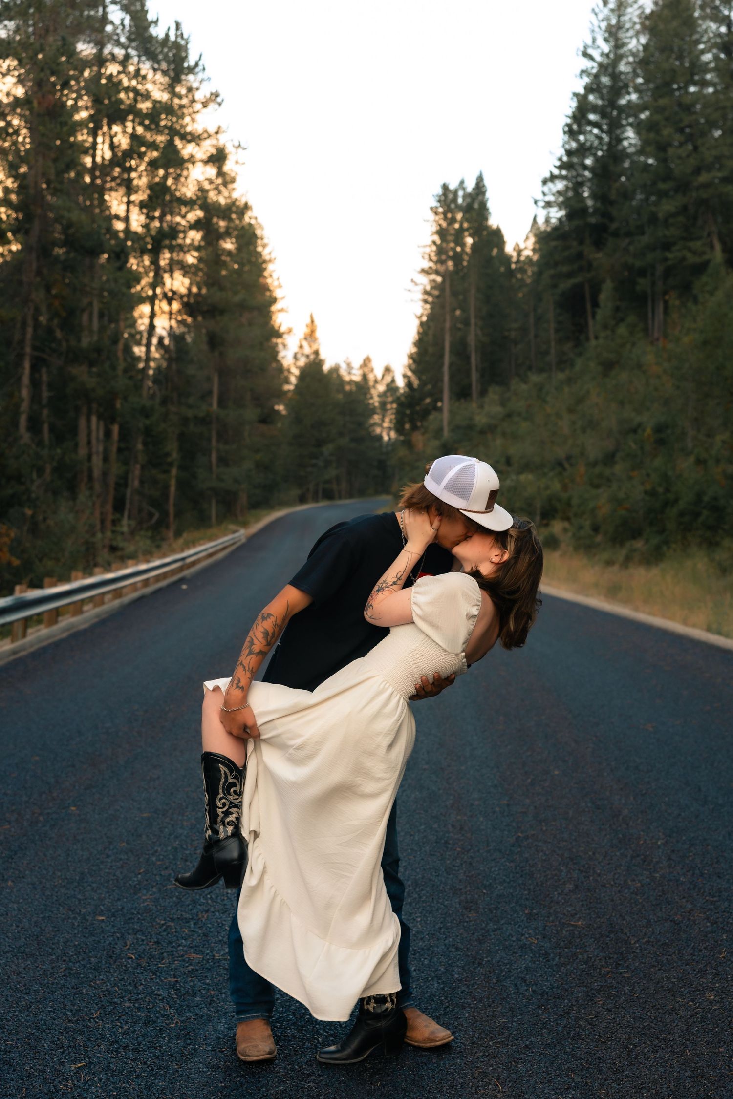 Couple sharing a romantic dip kiss on a forest road at sunset, woman in white dress and cowboy boots.