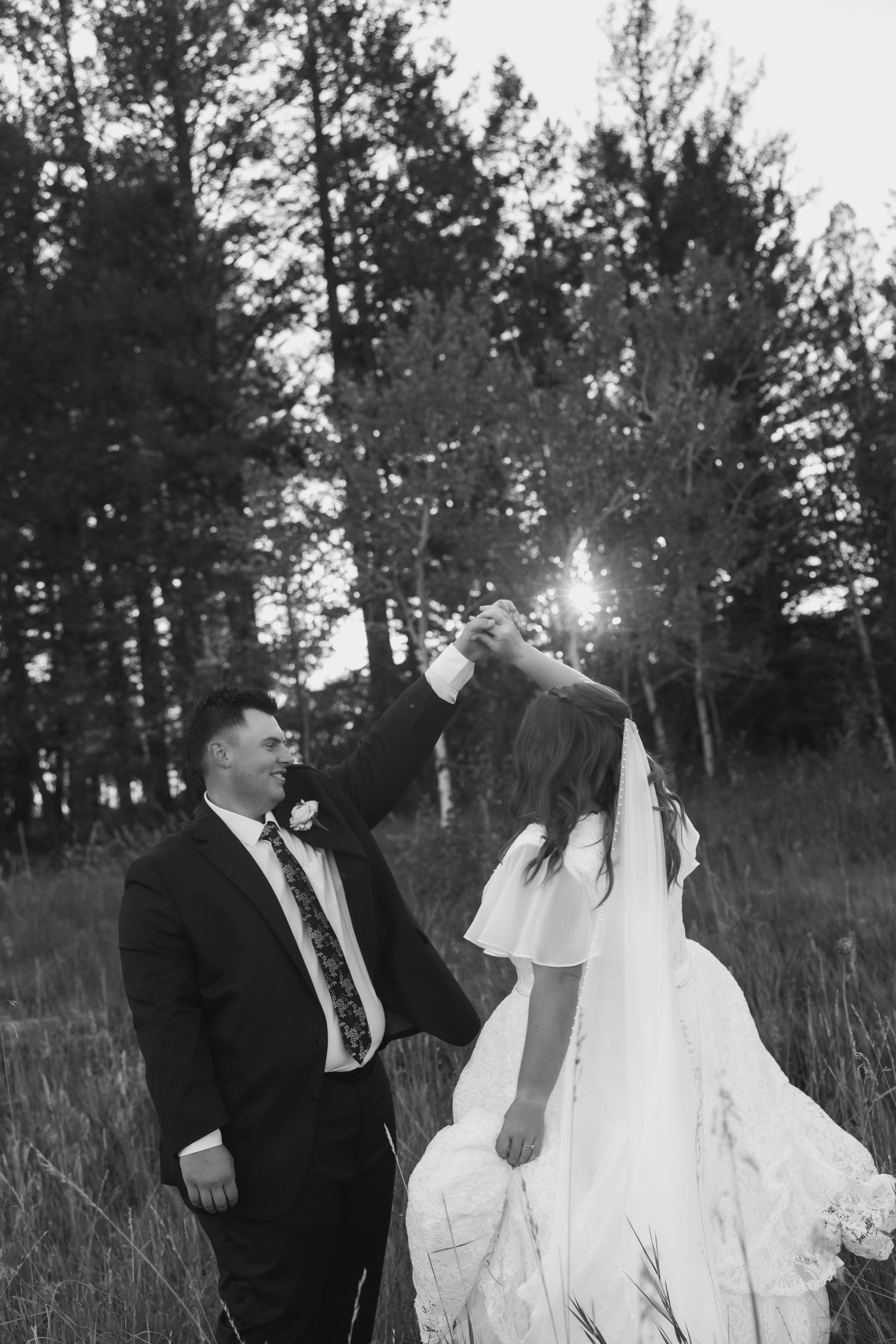 Black and white photo of a groom spinning his bride in a field surrounded by tall pine trees during golden hour.