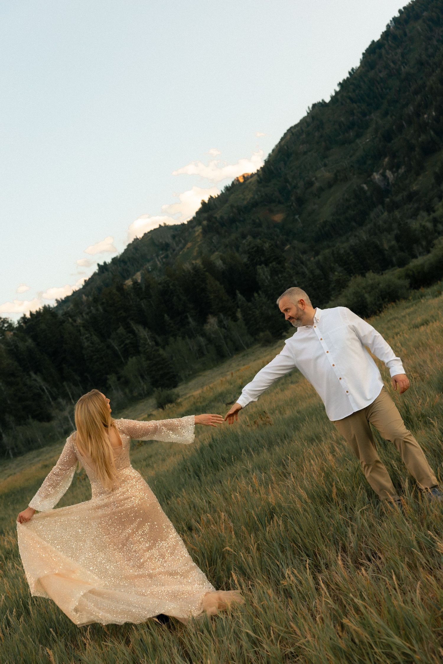 Couple dancing in a mountain meadow at dusk, woman in flowing gold gown, man in white shirt holding hands.
