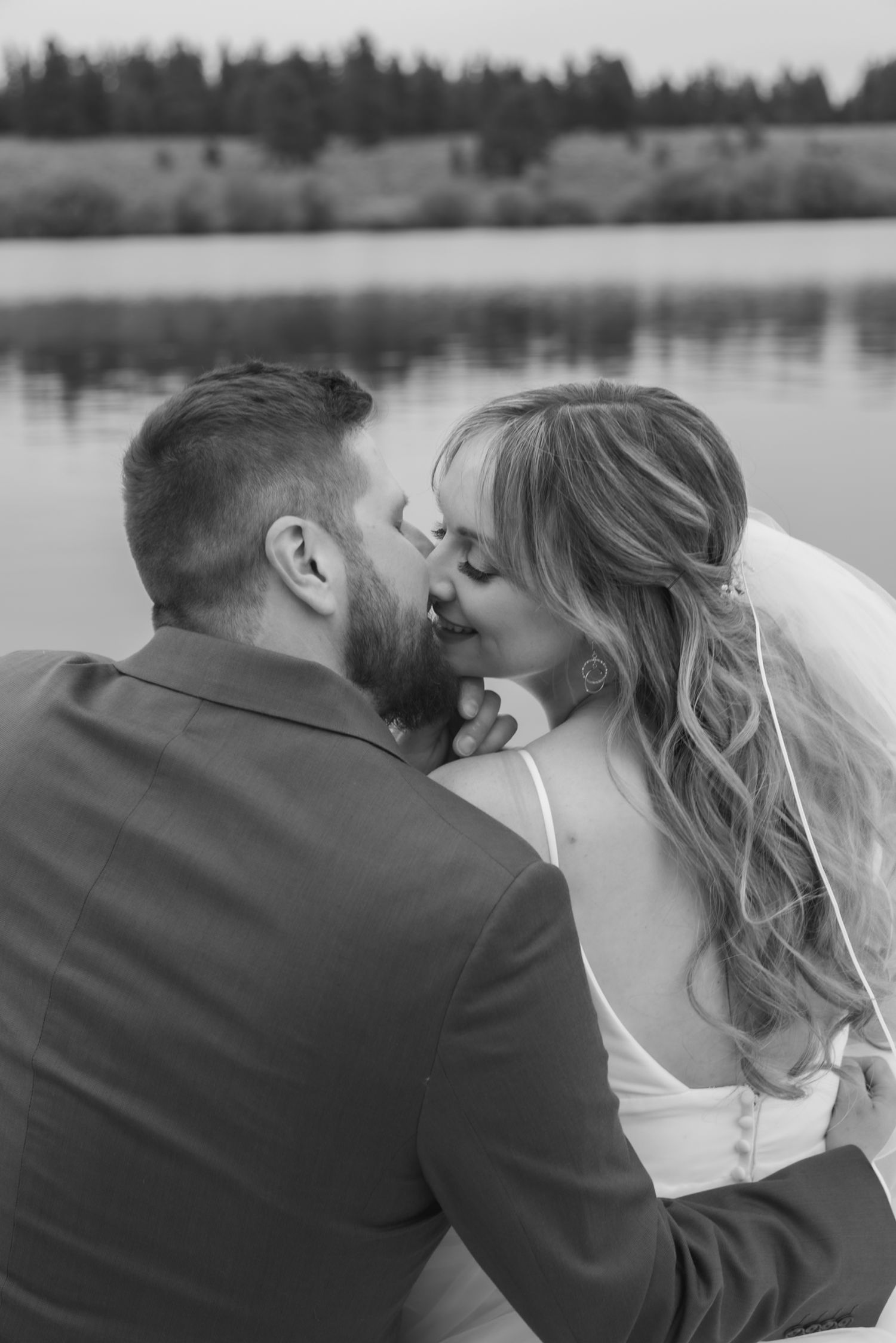 Black and white photo of a bride and groom sharing a romantic kiss by a serene lake on their wedding day.