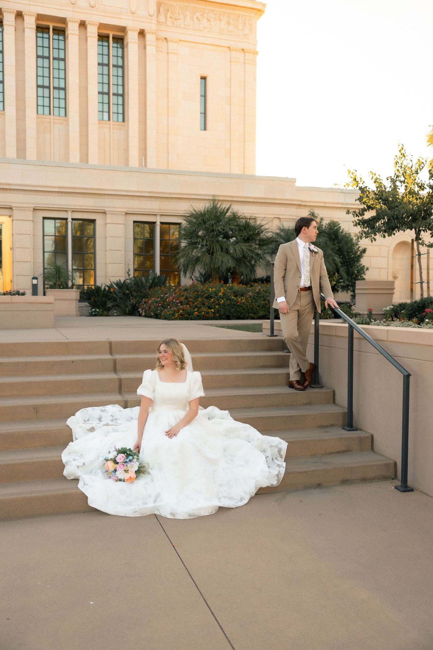 Bride in white gown sits on temple steps holding bouquet while groom in tan suit stands behind her.