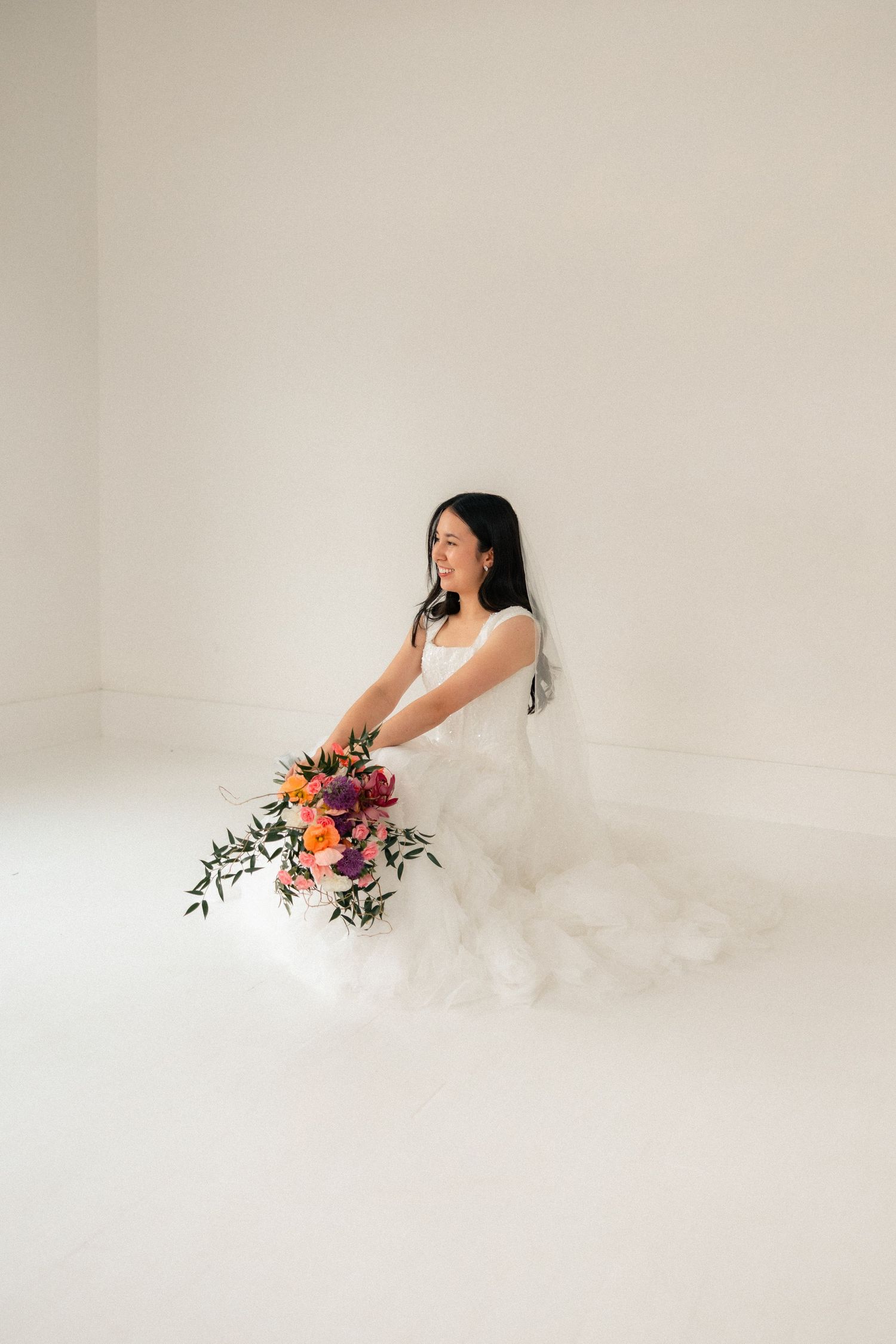 Bride in flowing white tulle wedding gown seated on floor holding vibrant orange and pink bridal bouquet.