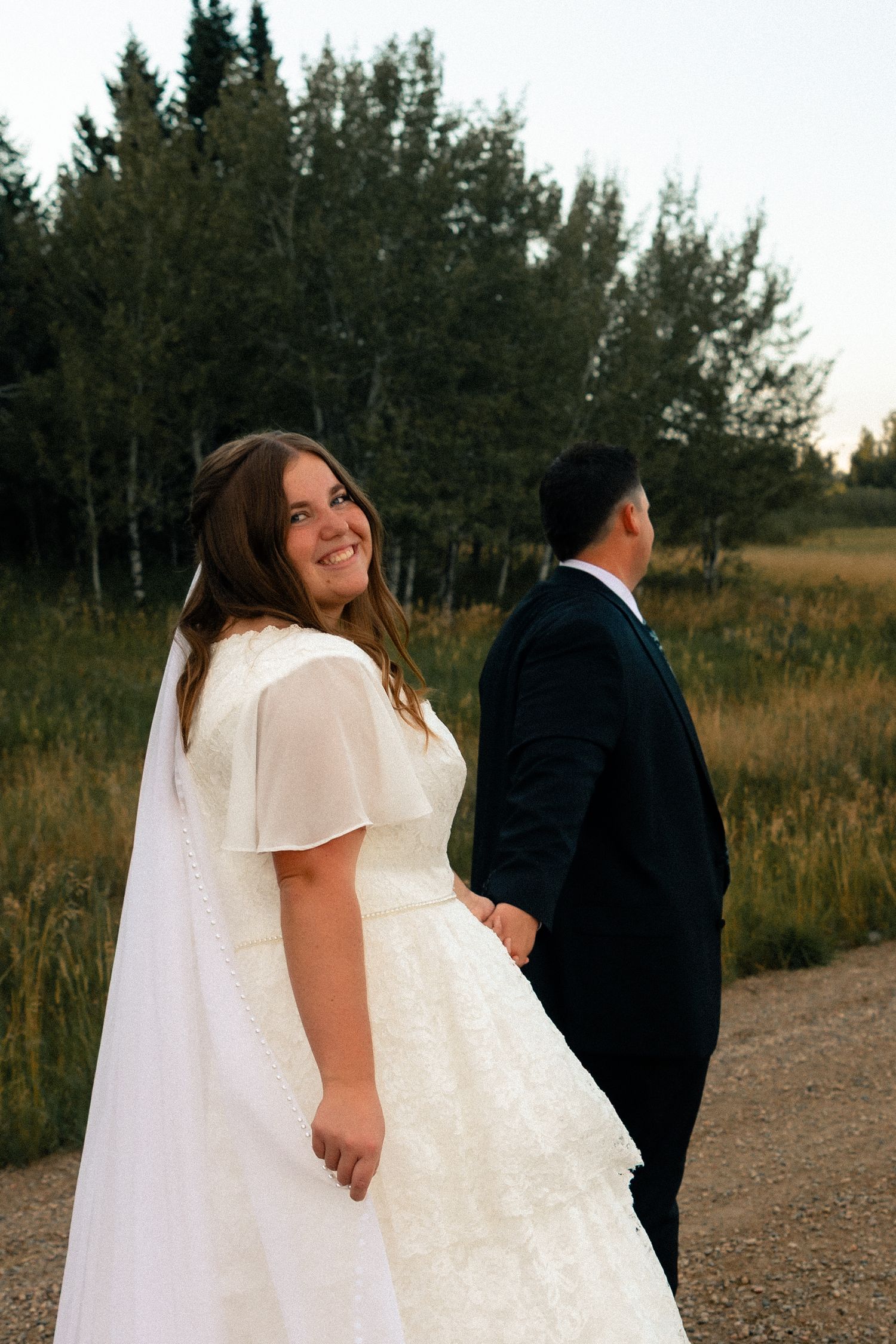 Bride in white lace gown smiles over shoulder while holding groom's hand on scenic outdoor path at golden hour.
