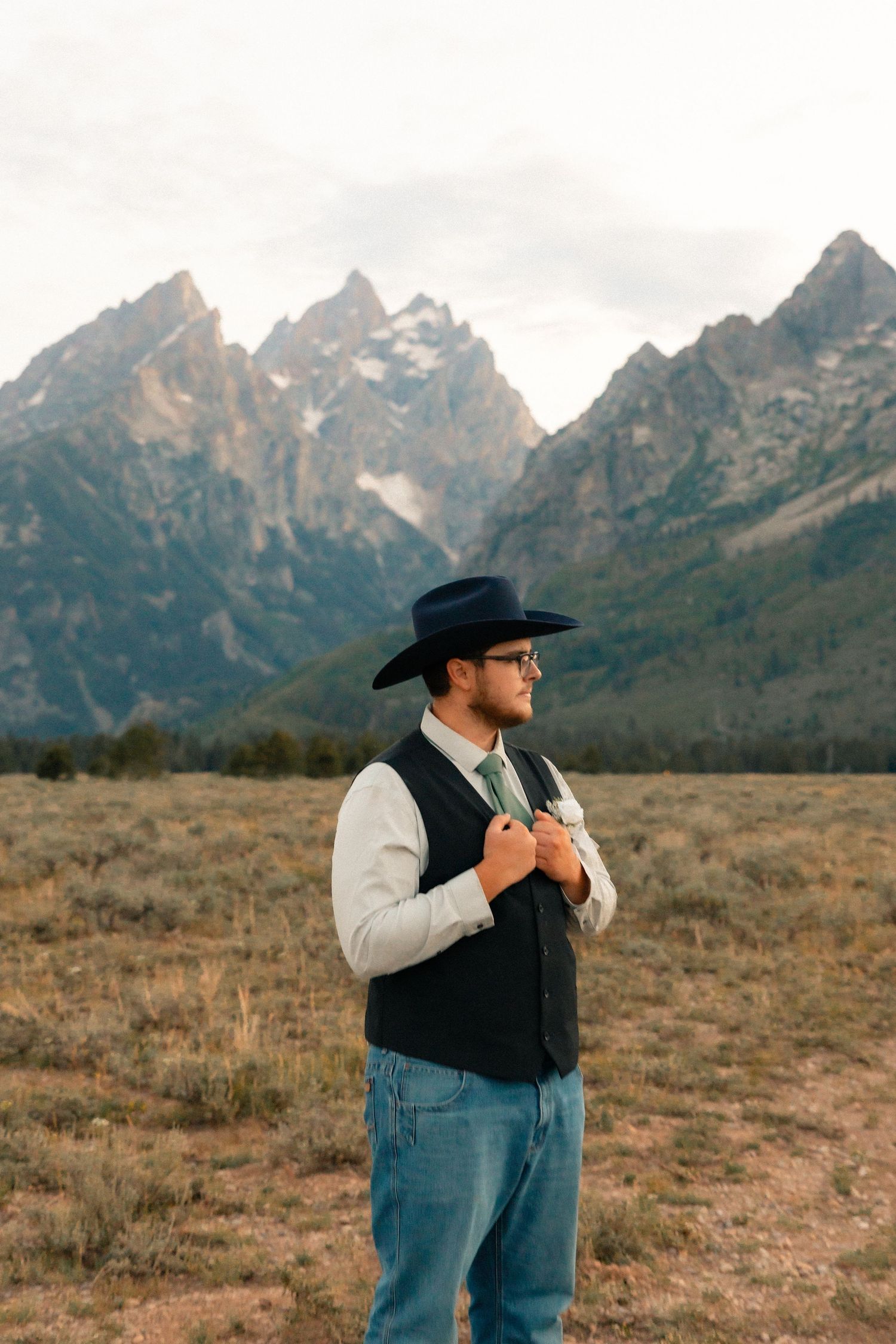 Groom in black cowboy hat and vest poses in field with Grand Teton mountains behind him.