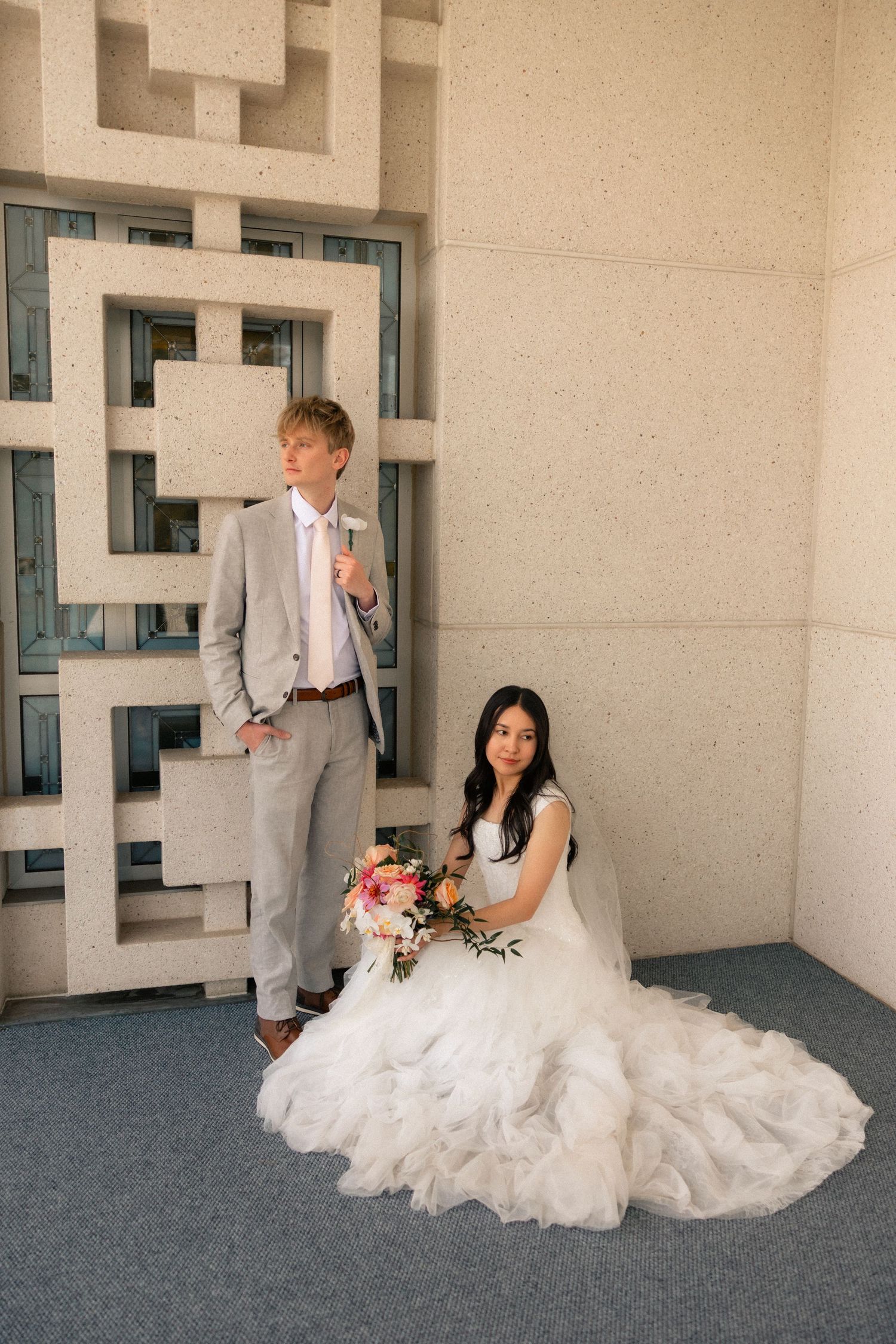 Bride in ruffled gown sits beside groom in gray suit near decorative stone wall during wedding portrait session.