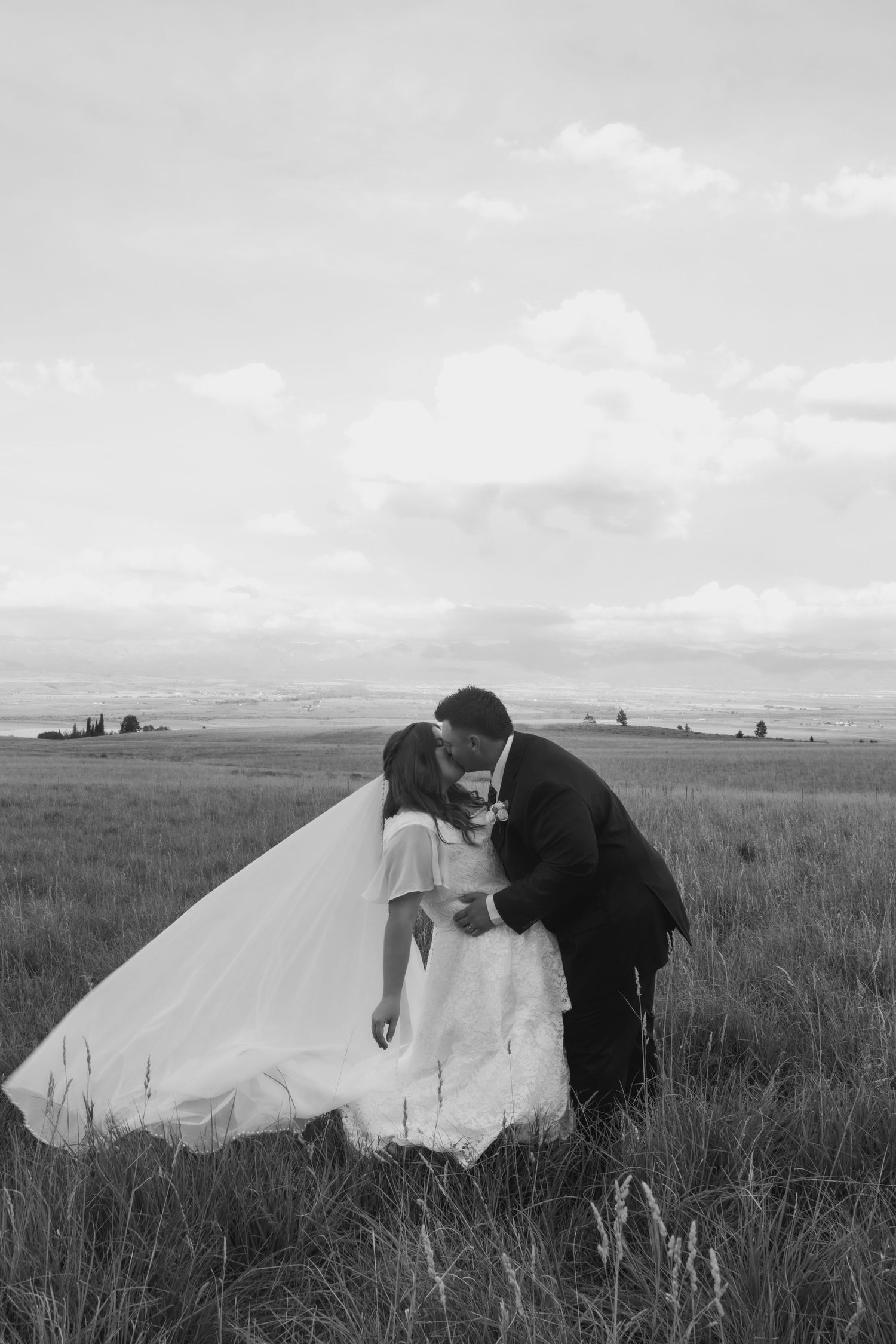 Black and white photo of a bride and groom sharing a romantic kiss in an open field with a dramatic cloudy sky.
