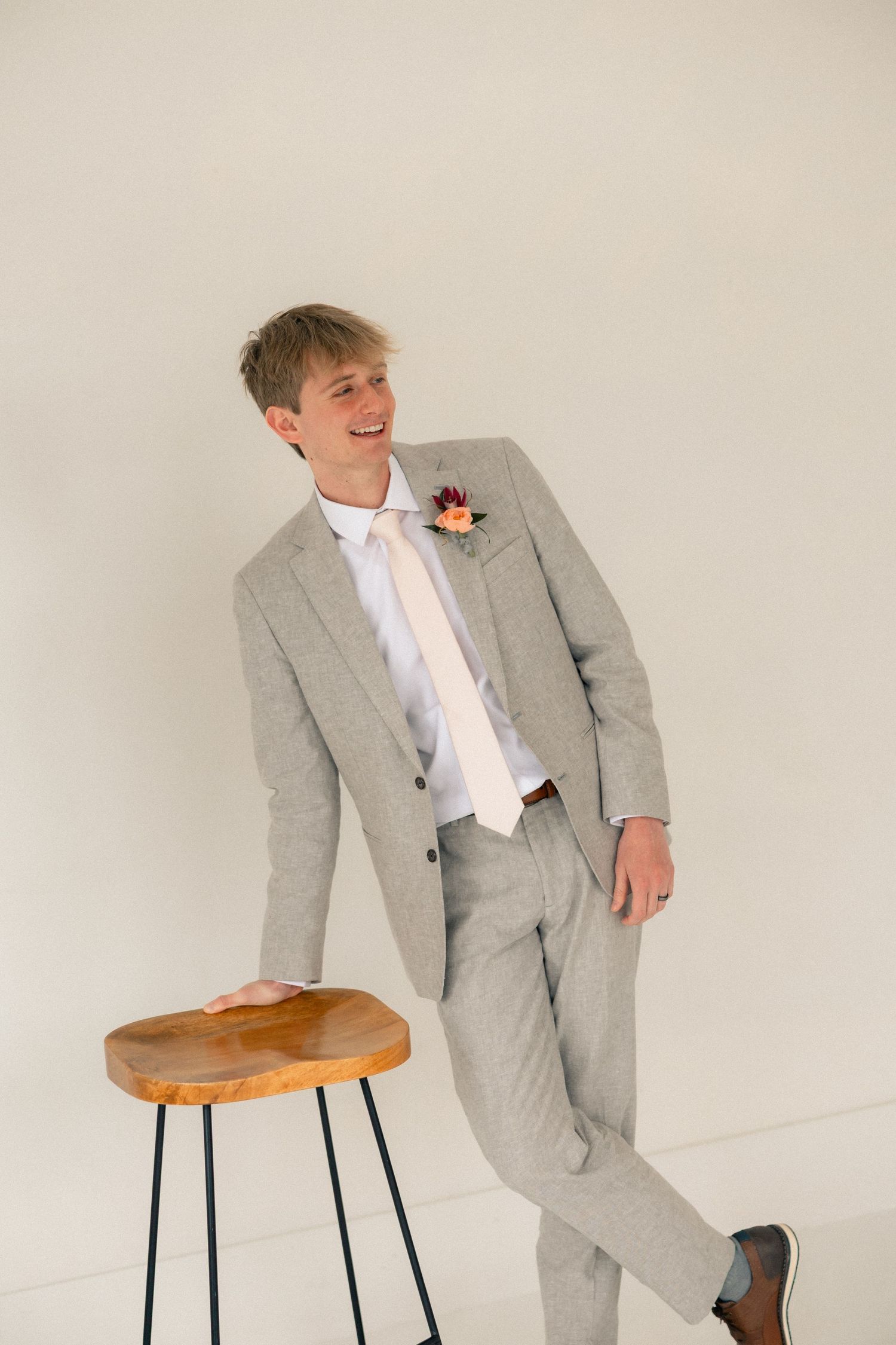 Smiling groom in light gray suit with floral boutonniere leaning on wooden stool against white background.