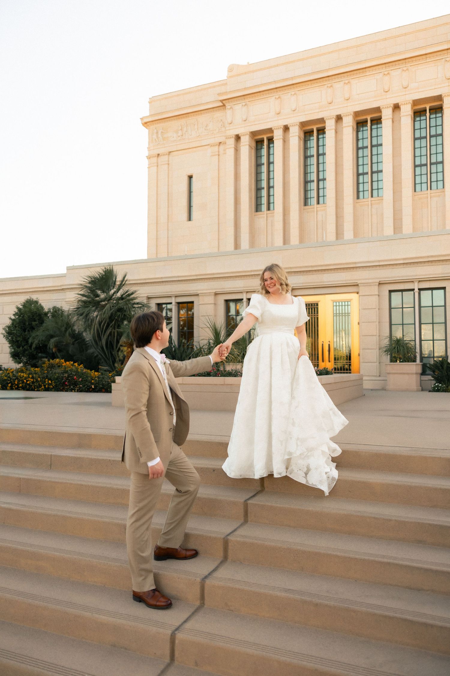 Bride and groom hold hands on temple steps, she in white gown, he in tan suit, classic columns behind them.