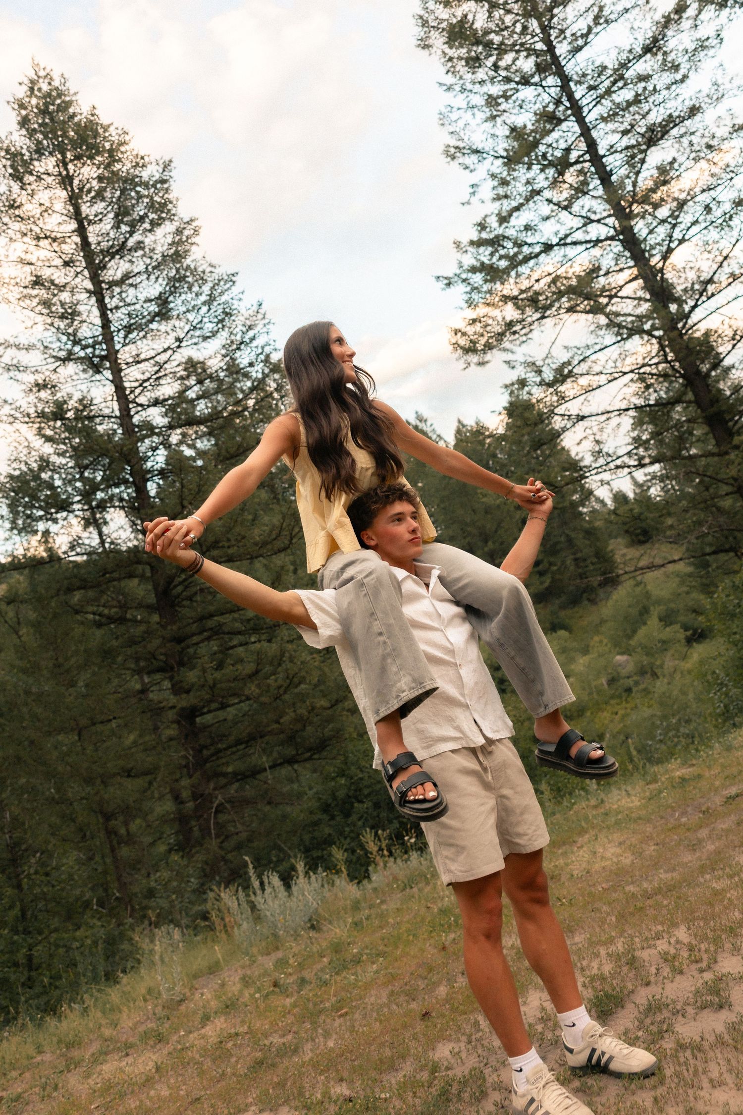 Young couple in forest, girl sitting on boyfriend's shoulders, holding hands, surrounded by tall pine trees.
