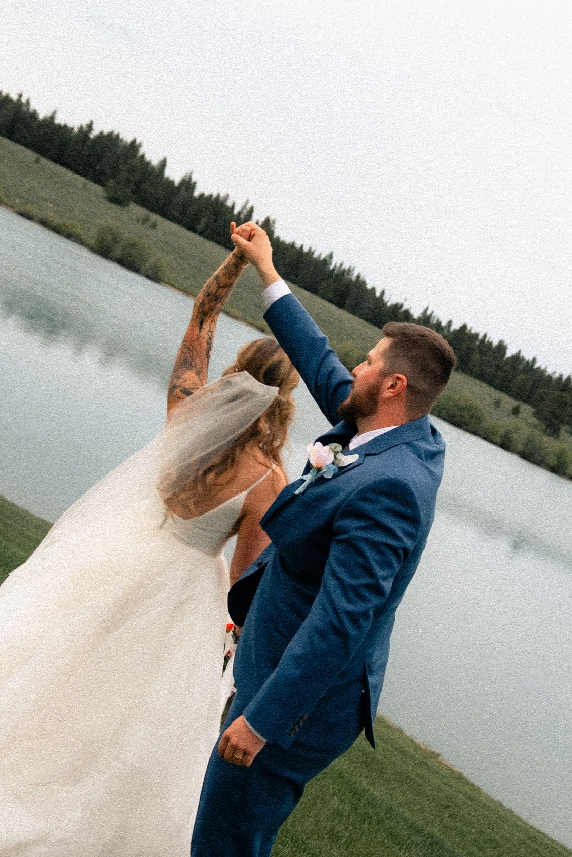 Groom in blue suit twirling bride in white wedding dress outdoors by a lake with green trees in background.