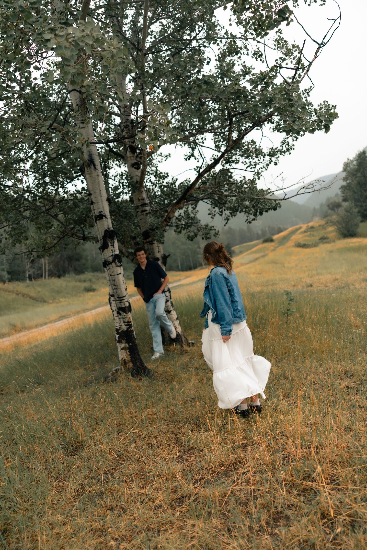 Couple in a meadow near birch trees, woman in white skirt and denim jacket walks toward man in casual attire.
