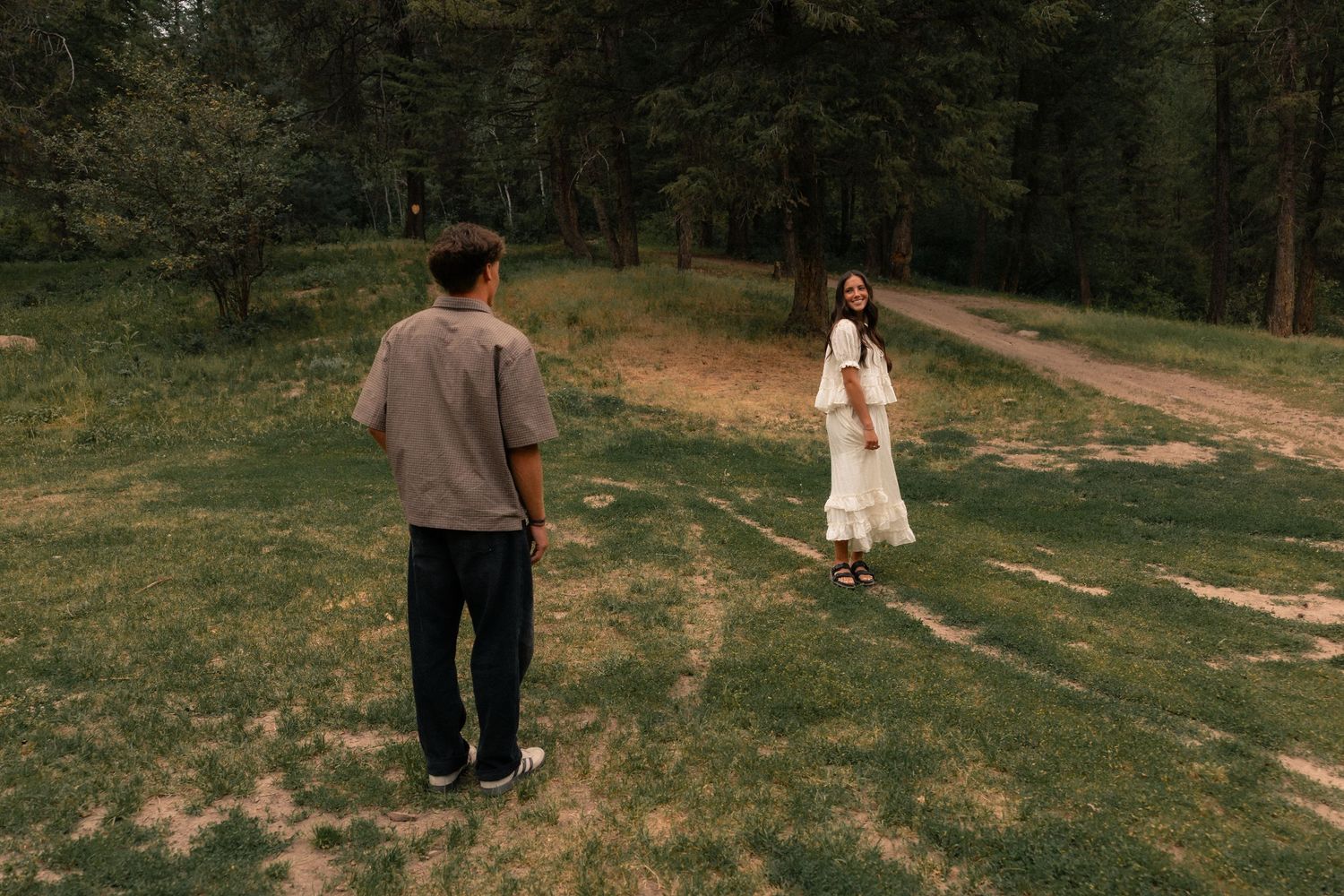 A couple stands apart on a grassy forest clearing, woman in white dress facing man in casual gray outfit.