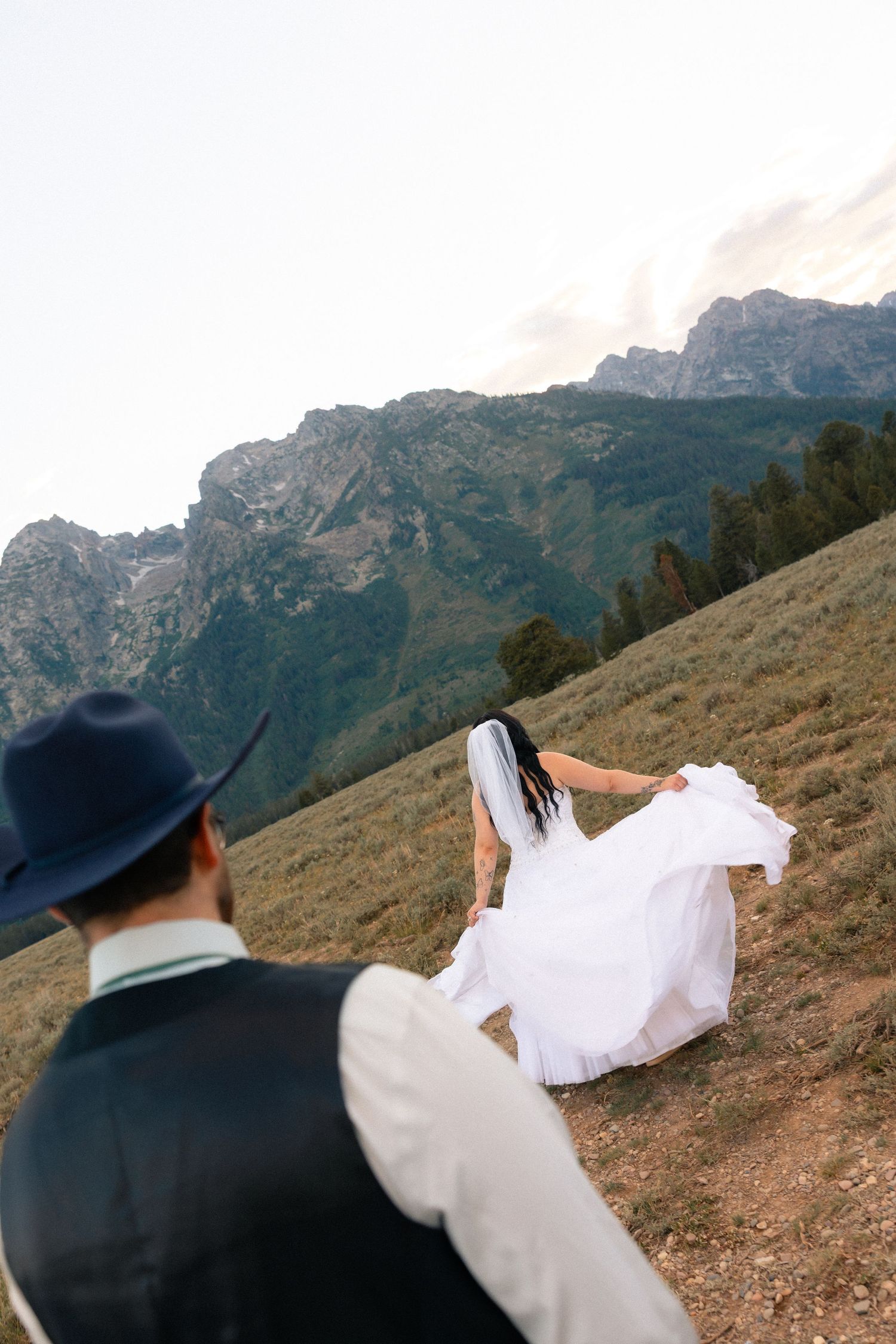 Bride in white dress and groom in cowboy hat walk toward mountain landscape during outdoor wedding photoshoot.