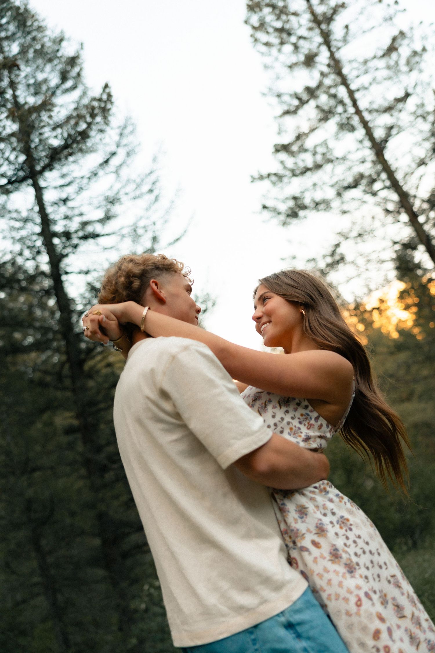 A joyful young couple embraces and smiles at each other in a forest at sunset.