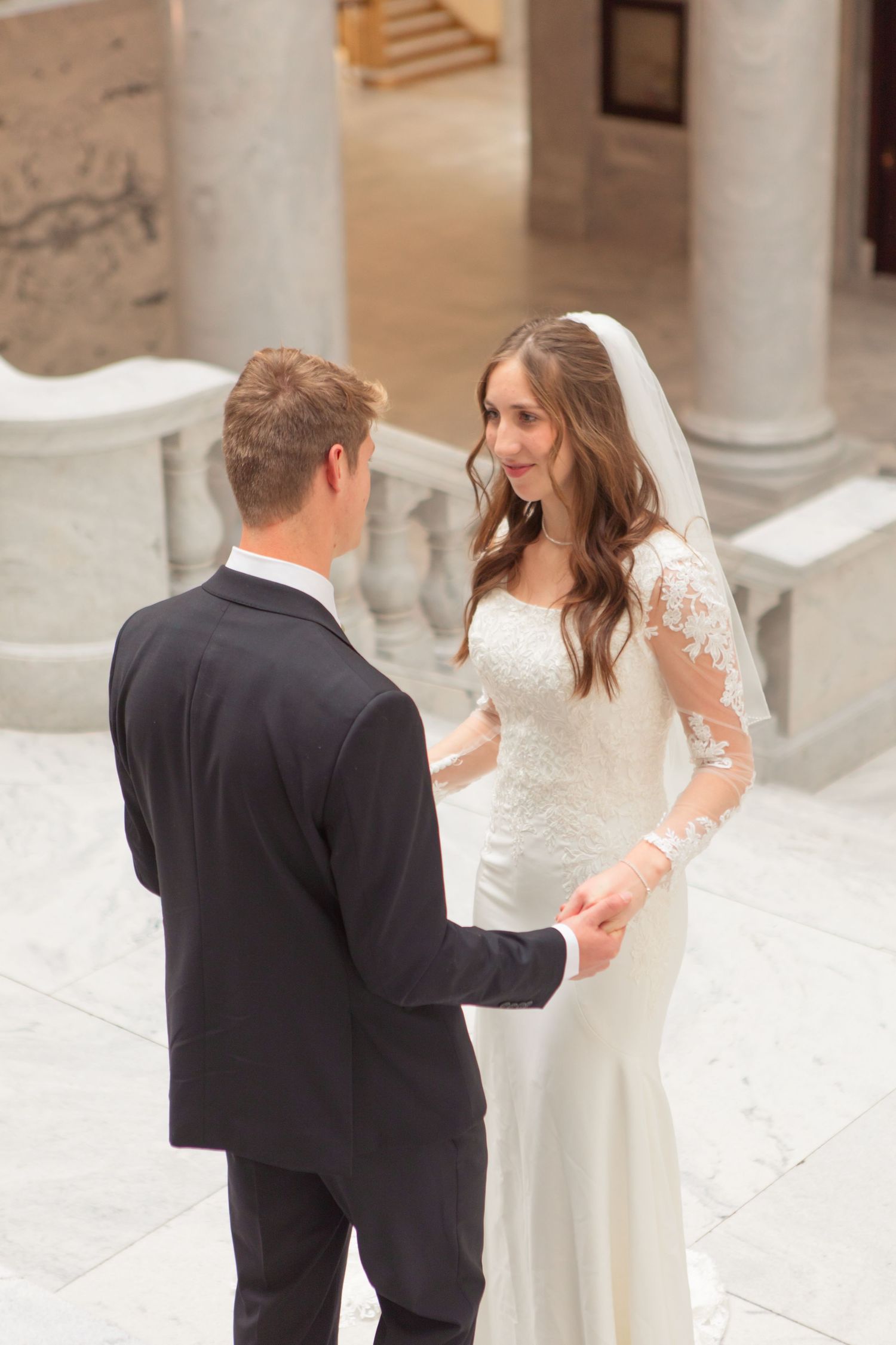 Bride and groom holding hands during wedding ceremony in elegant marble venue, bride wearing lace long-sleeve gown and veil.