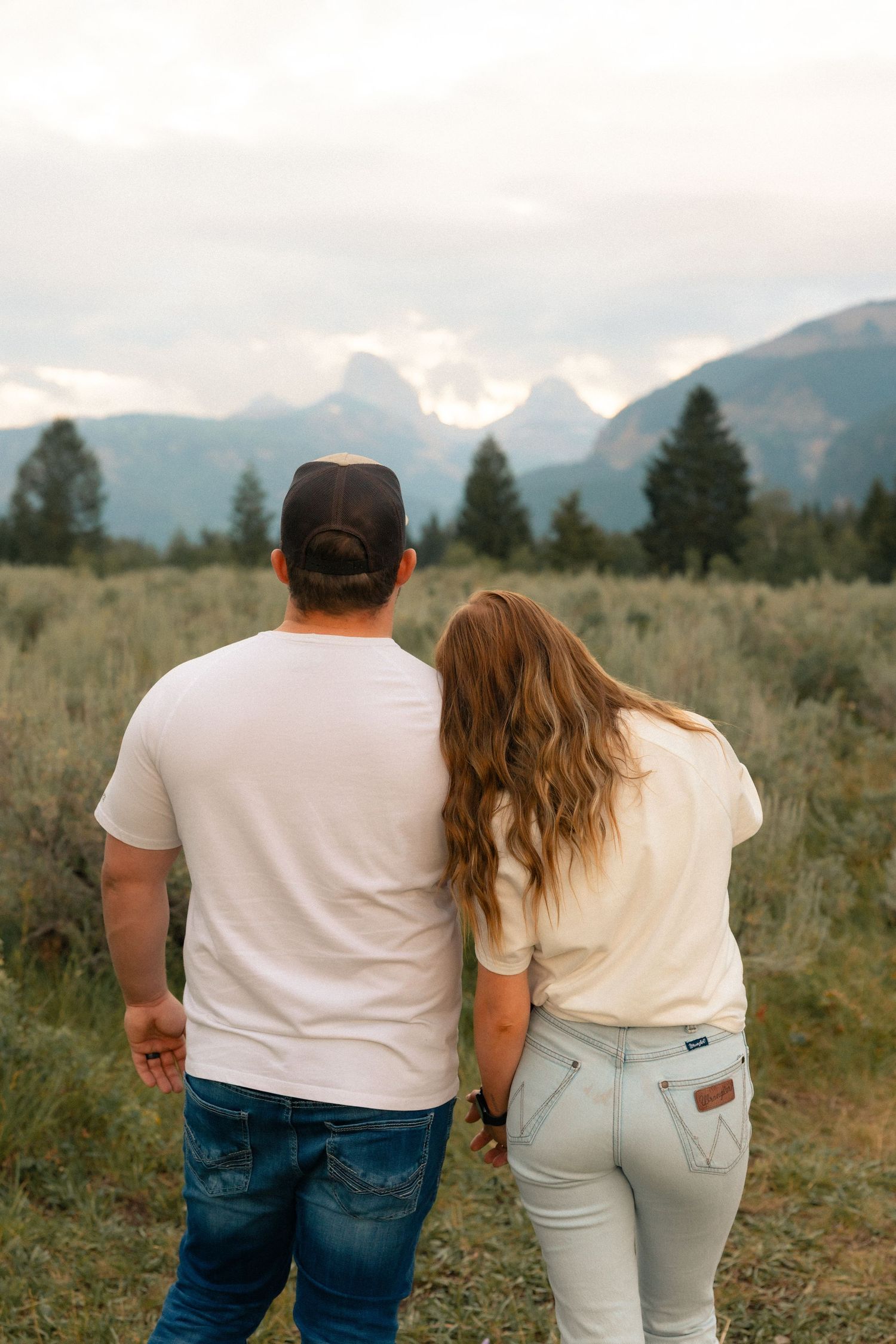 Couple holding hands gazing at Grand Teton mountain range surrounded by alpine meadows and evergreen trees.