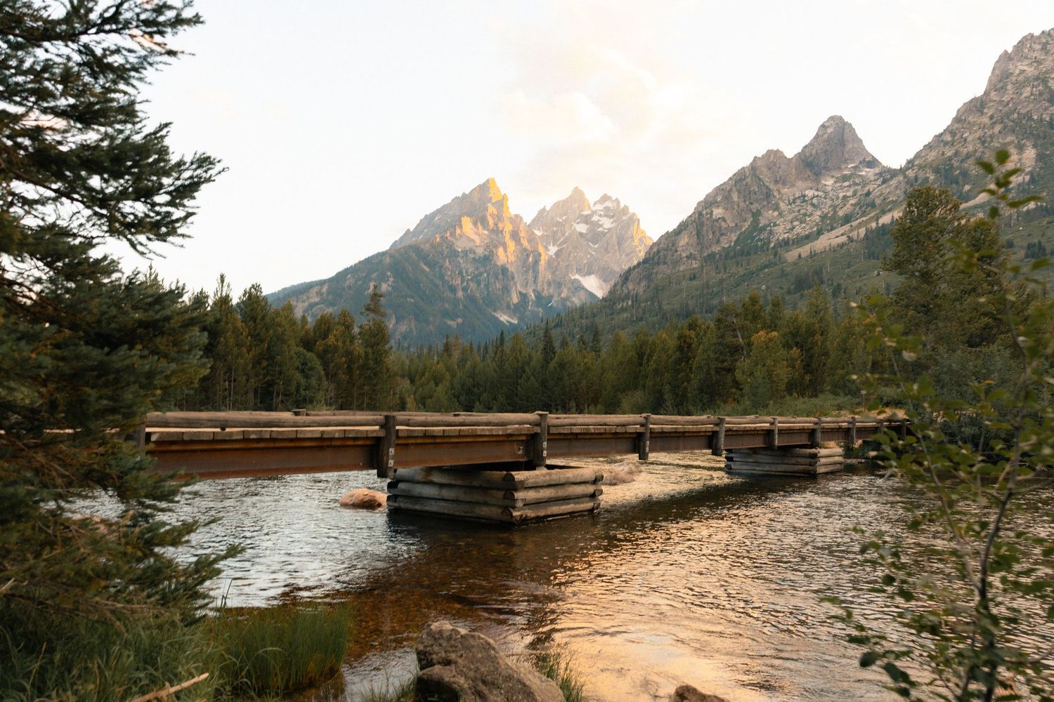 Wooden bridge over a calm river with Grand Teton mountain peaks and pine forest in the background at sunset.