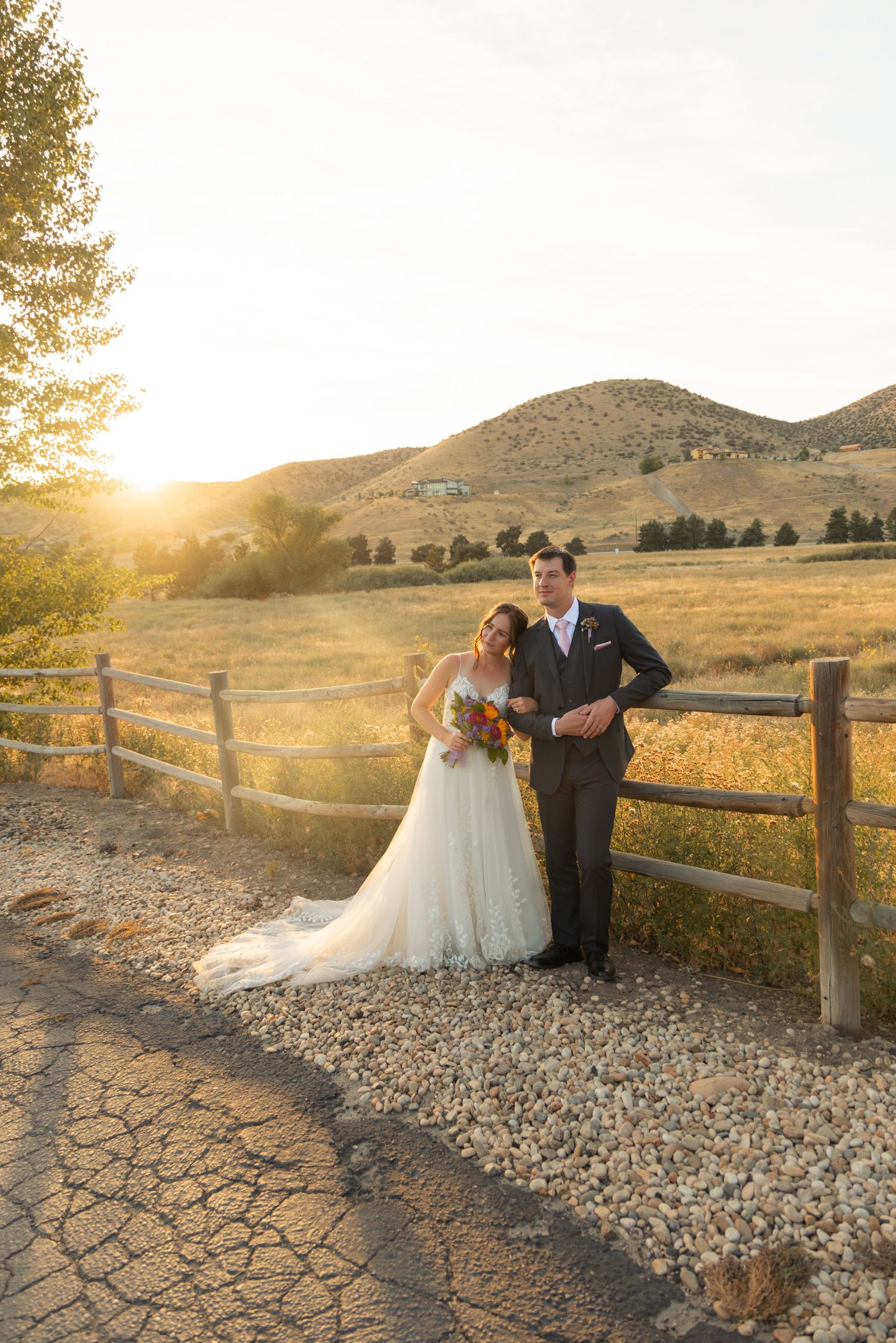 Bride and groom pose by rustic fence at golden sunset with rolling hills backdrop on their wedding day.