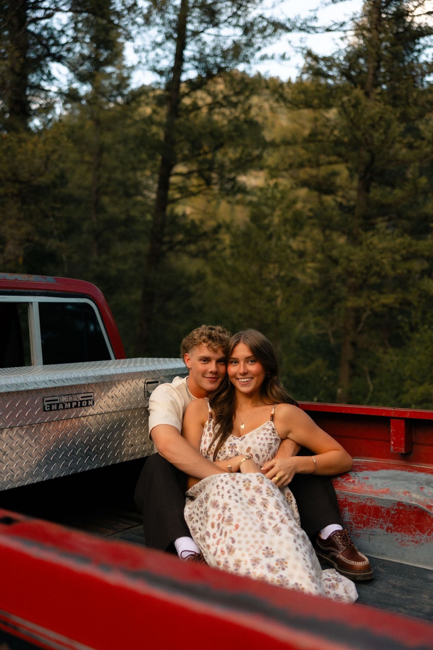 Young couple sitting together smiling in the bed of a red pickup truck surrounded by lush green forest at sunset.
