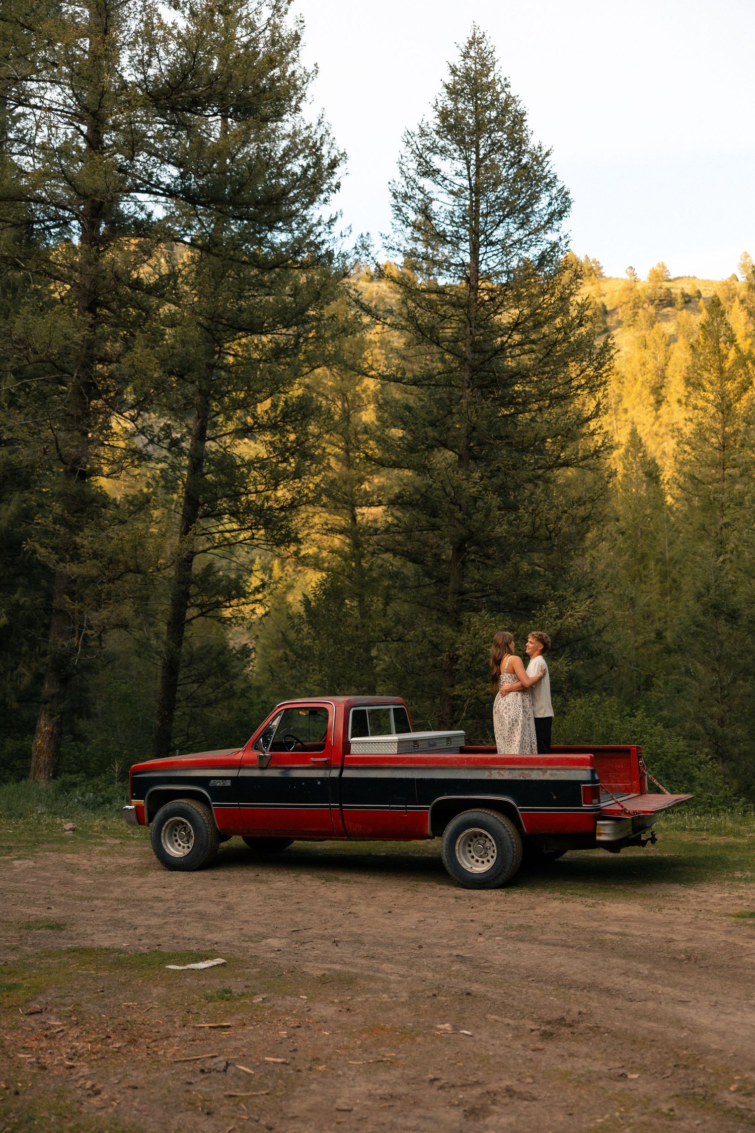 Couple in wedding attire standing in red pickup truck bed surrounded by tall pine trees and golden mountain backdrop.