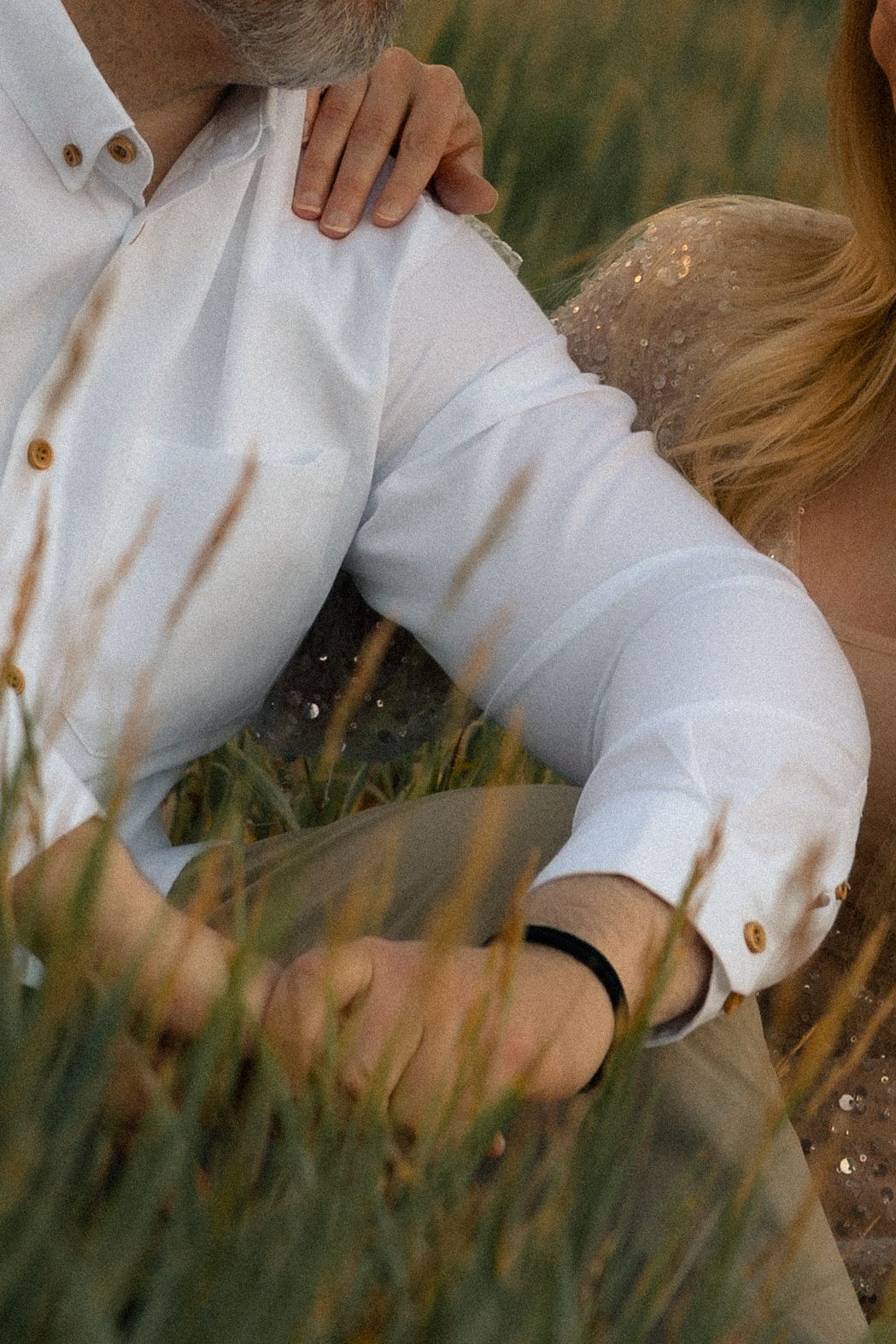Couple embracing in a field, man in white linen shirt with black bracelet, woman in lace dress resting head on his shoulder.