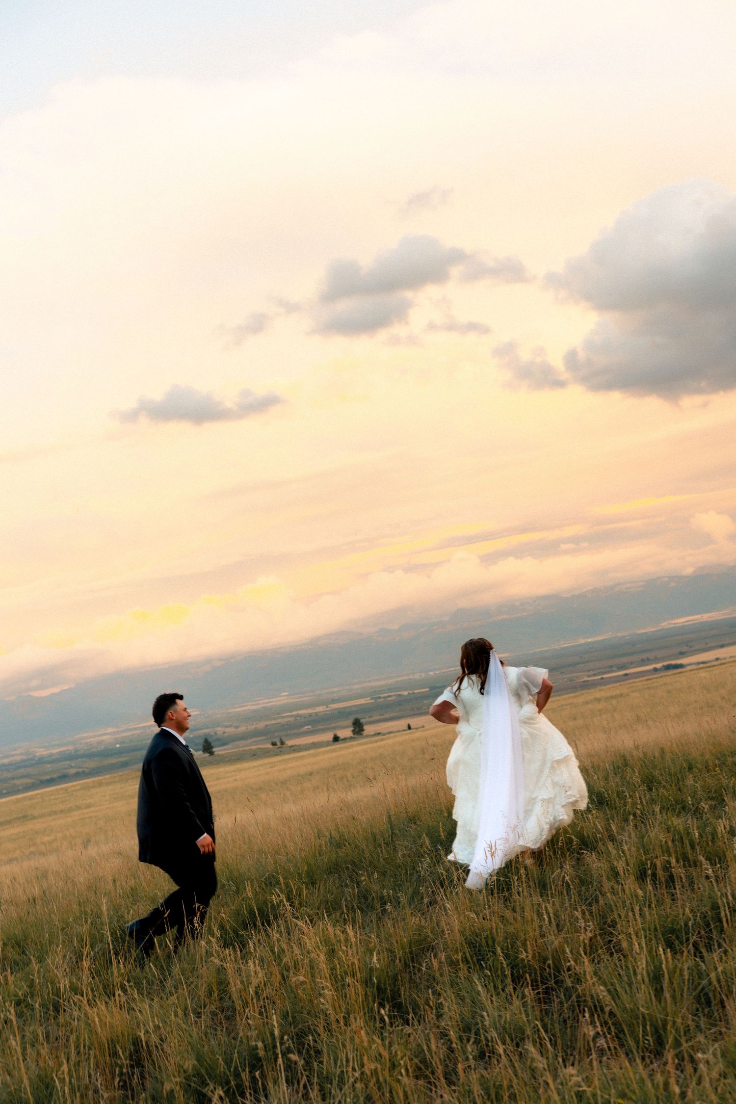 Bride and groom walking through golden field at sunset, bride in white gown with veil, groom in black tuxedo.