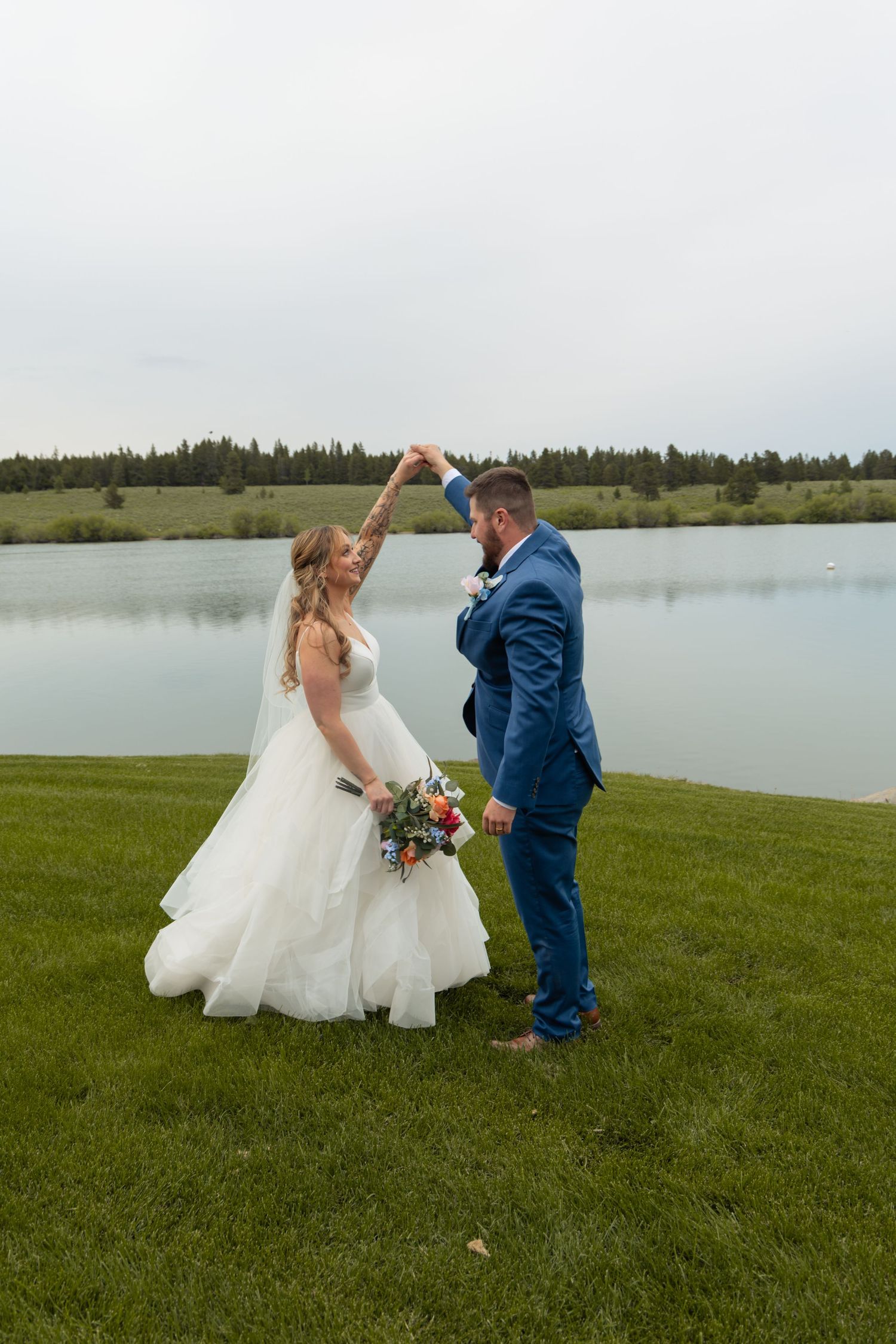 Bride in white gown and groom in blue suit dancing by a serene lake on lush green grass on their wedding day.