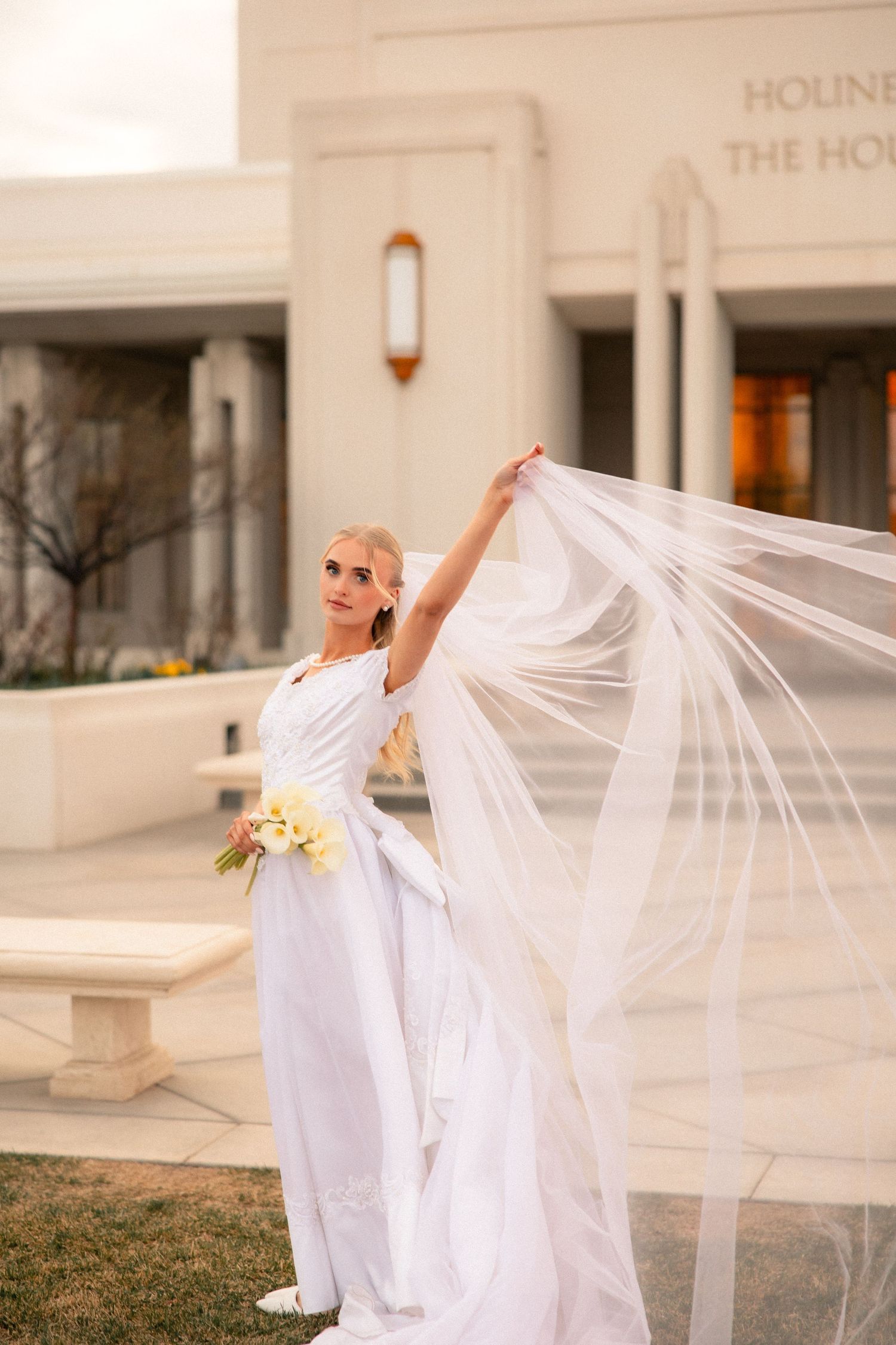 Bride in white gown holds flowing veil aloft outside a temple, holding a bouquet of white flowers.
