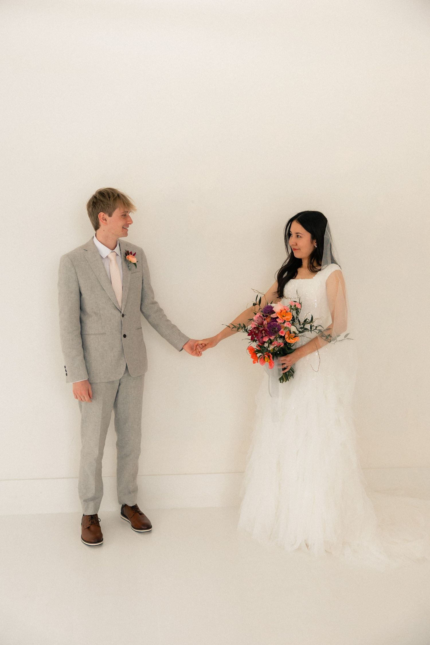 Bride and groom holding hands in white studio, she wears white gown with colorful bouquet, he wears gray suit.