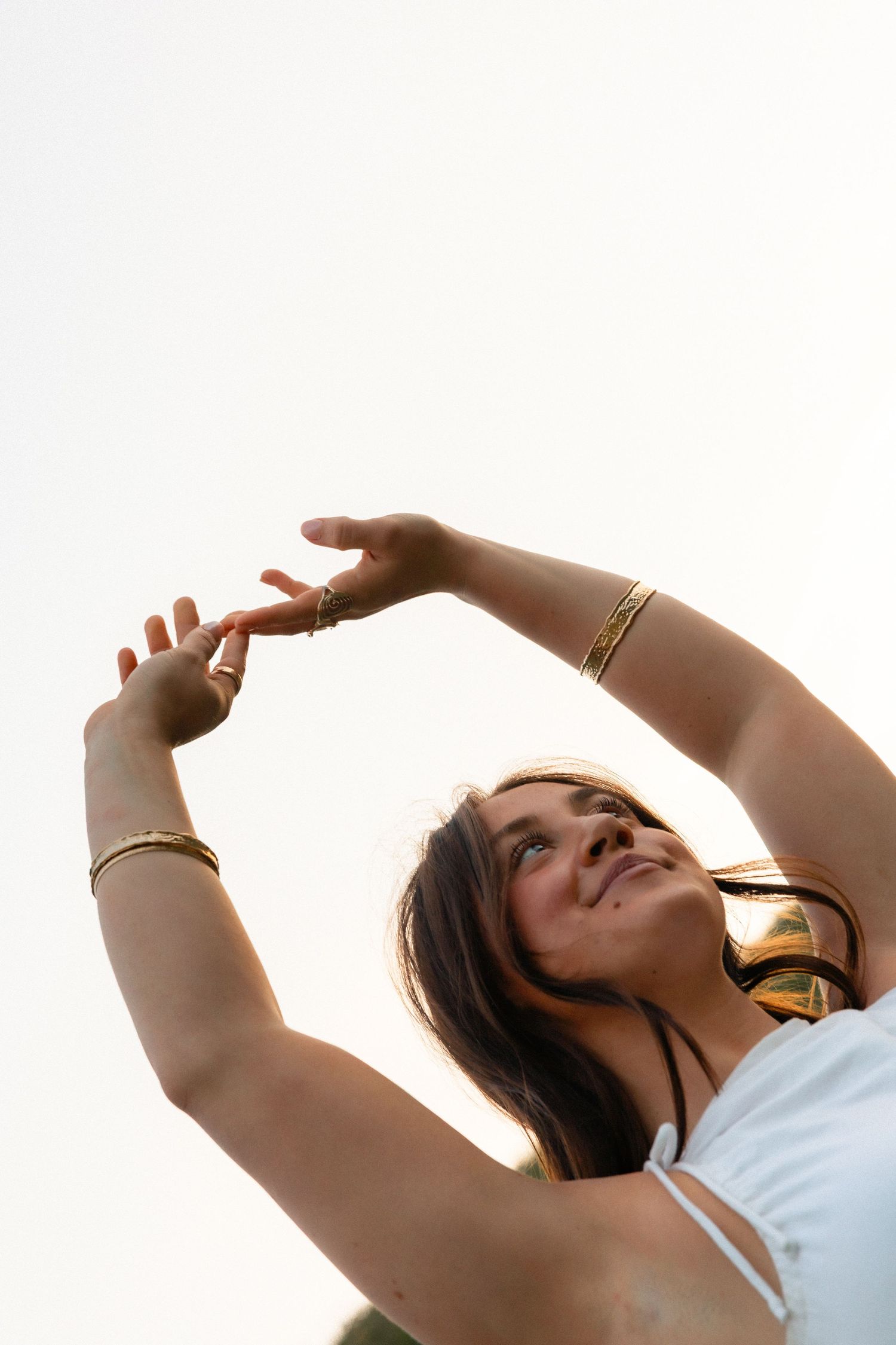 Young woman with gold bracelets and headphones raises arms gracefully against bright sky backdrop.