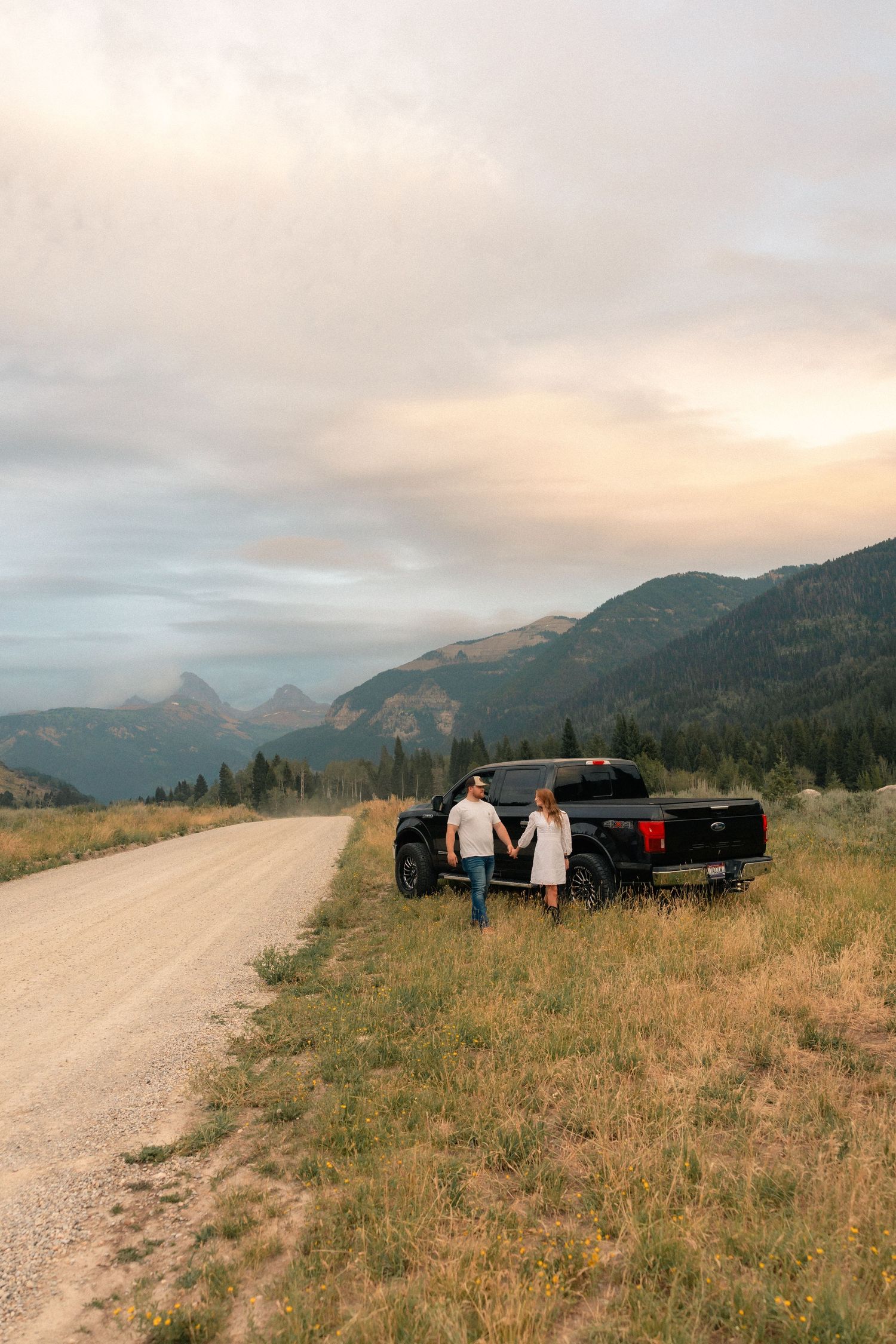 Couple holding hands by black pickup truck on gravel road with mountain sunset backdrop.