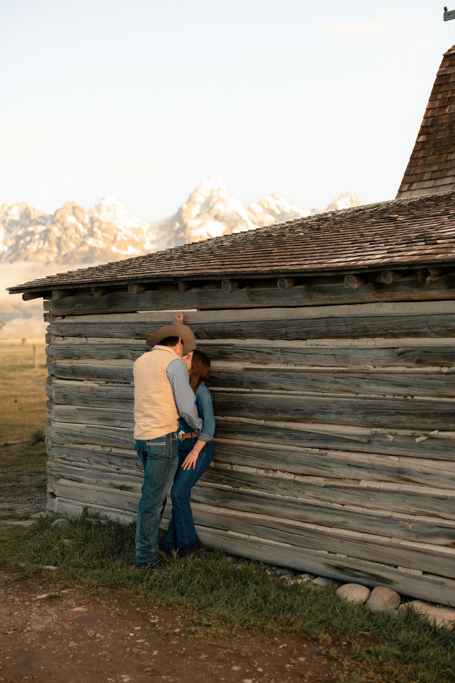 Couple sharing a romantic moment leaning against a rustic log cabin with Grand Teton mountains in the background.