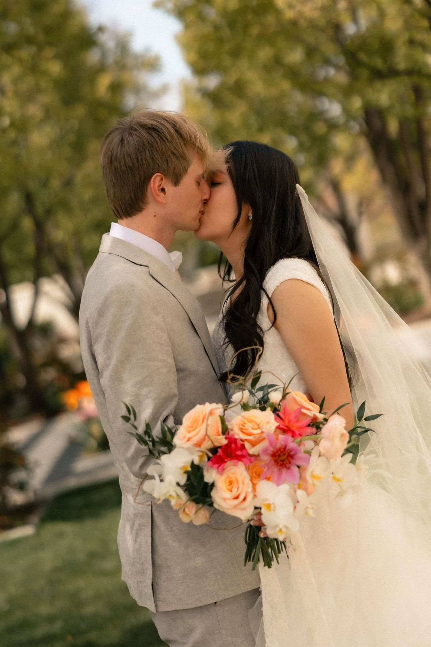 Bride and groom share a kiss outdoors, bride holding a colorful bouquet of peach roses and pink flowers, groom in gray suit.