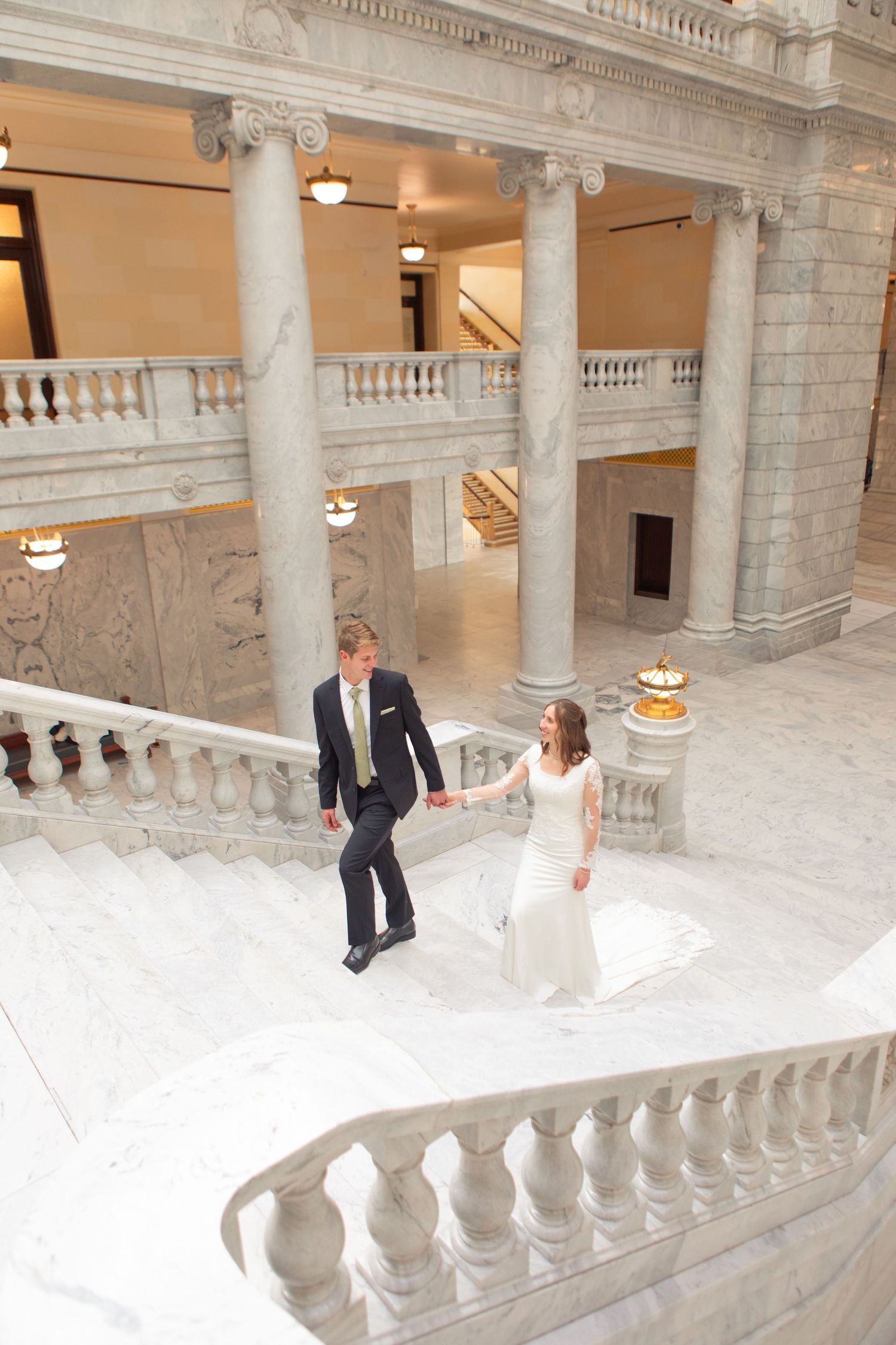 Bride and groom holding hands walking up grand marble staircase in elegant classical building.