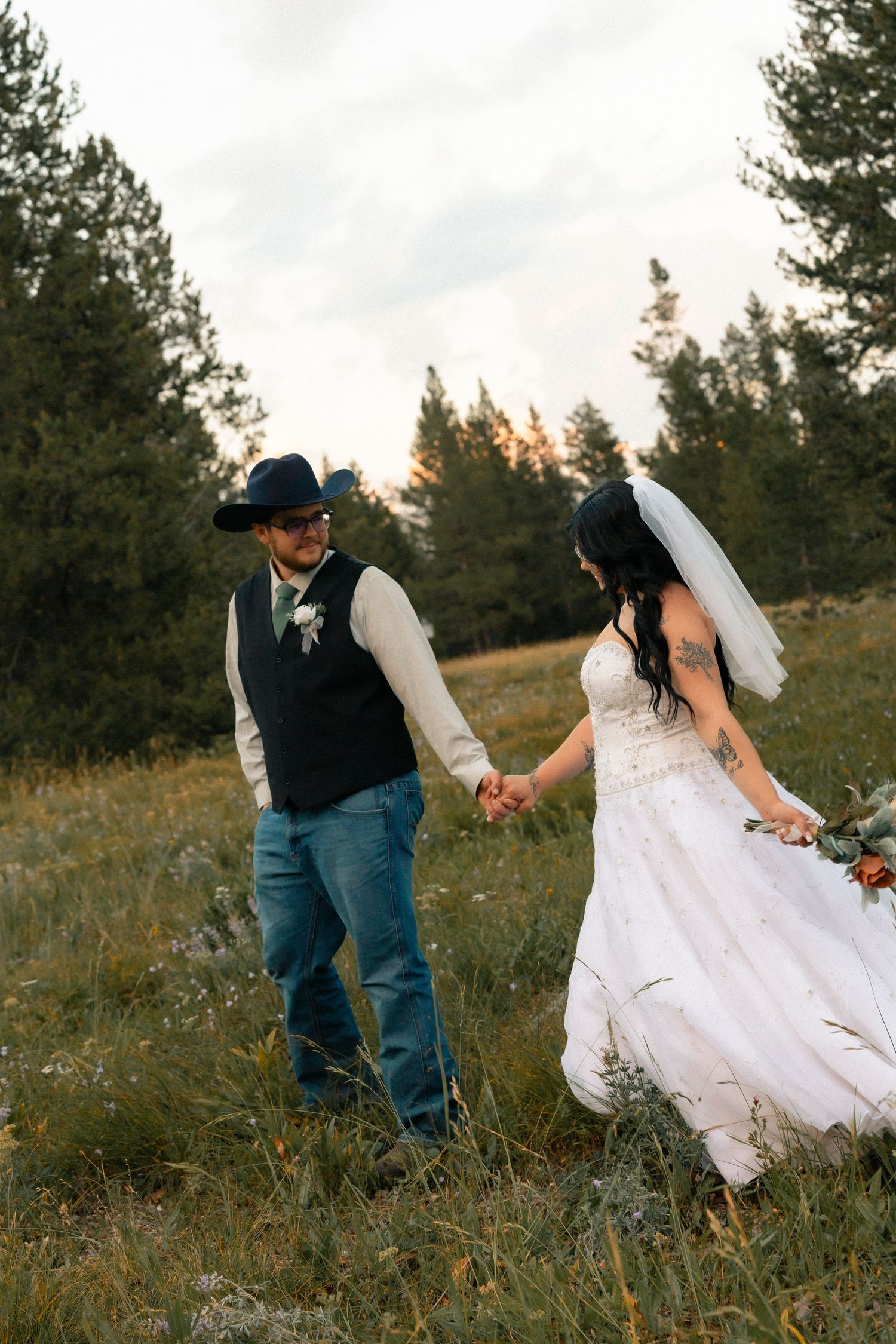 Bride and groom hold hands walking through a meadow, groom in cowboy hat and jeans, bride in white gown with veil.