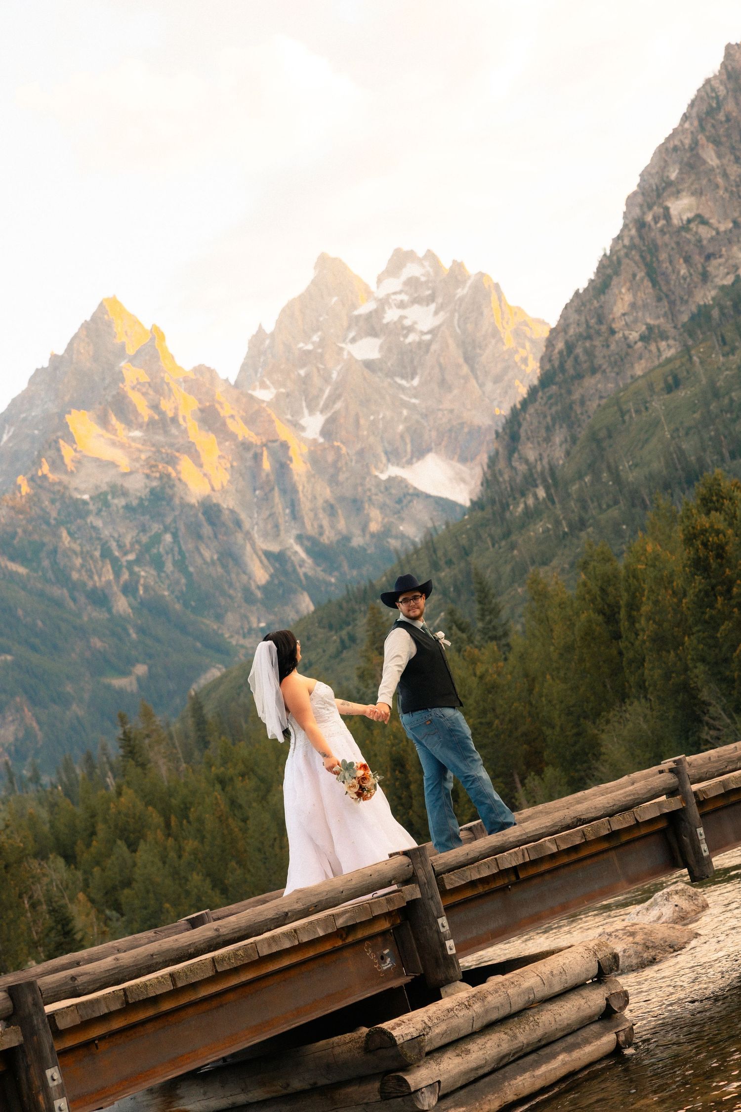 Bride and groom hold hands on a rustic wooden bridge with the Grand Teton mountains glowing at sunset behind them.