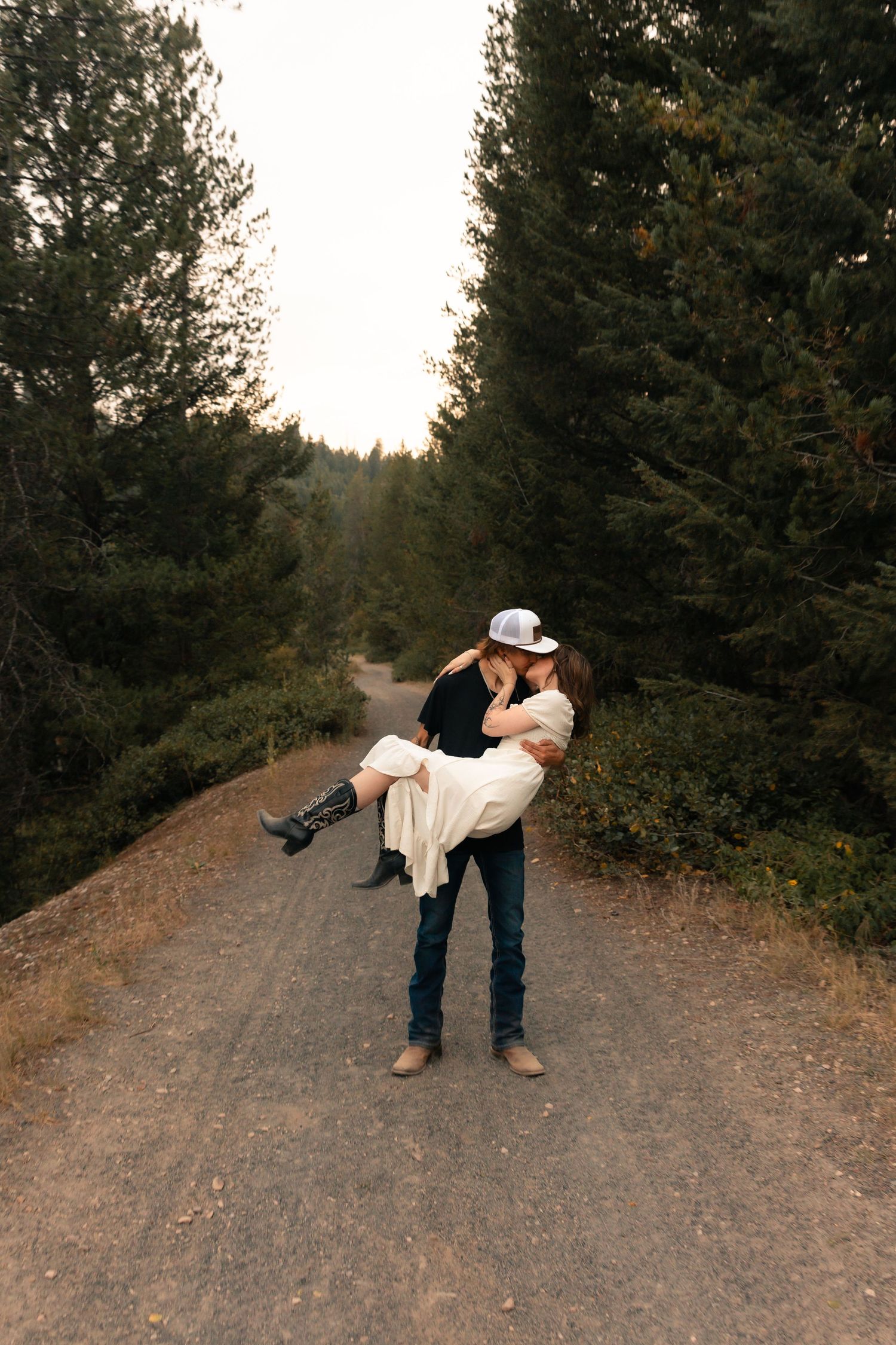 A couple shares a romantic kiss on a forest dirt road, man carrying woman in white dress and cowboy boots.