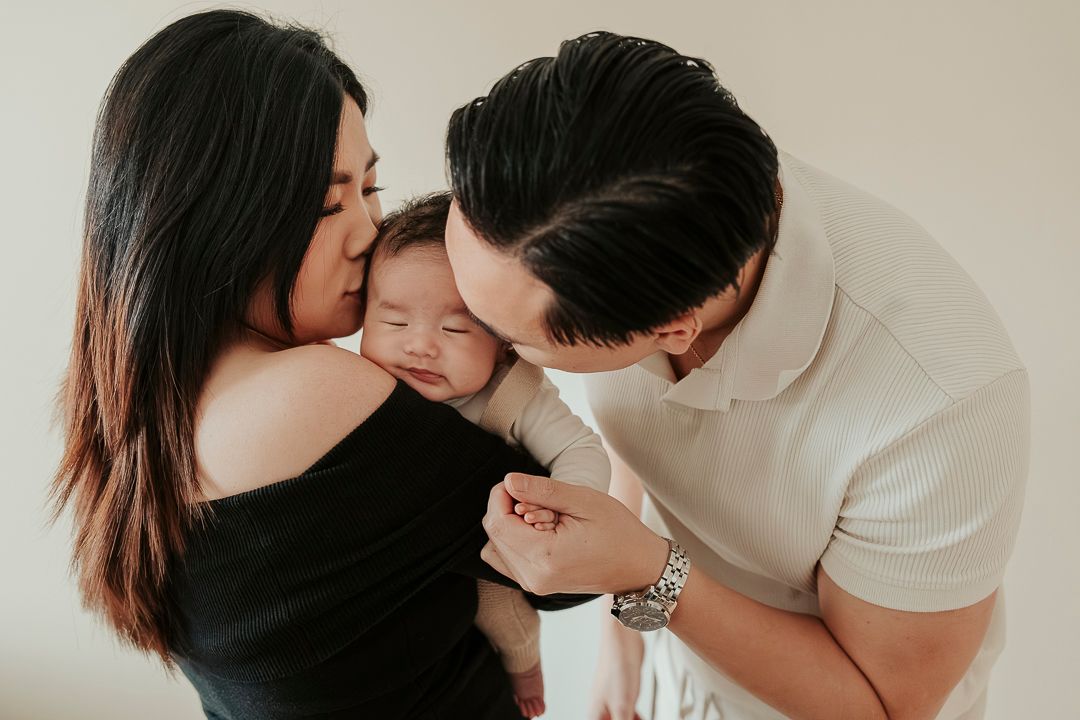 En familjefotografering i hemmet i Göteborg. Mamman har barnet i famnen och båda föräldrarna pussar på barnets huvud.