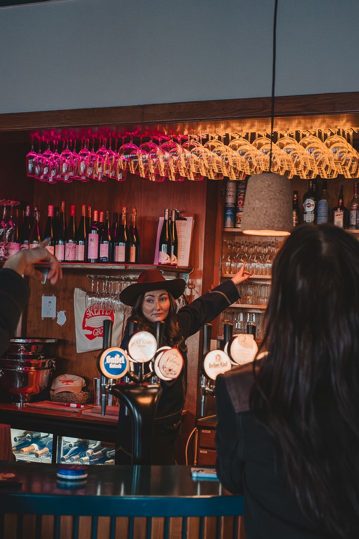 En kvinna i cowboy hat som står bakom baren och pekar på ett glas. En annan kvinna sitter med ryggen mot kameran