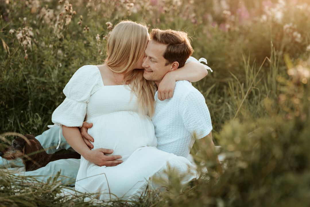 Par som sitter på en äng i golden hour. Kvinnan är gravid och sitter i mannens knä och de håller om varandra
