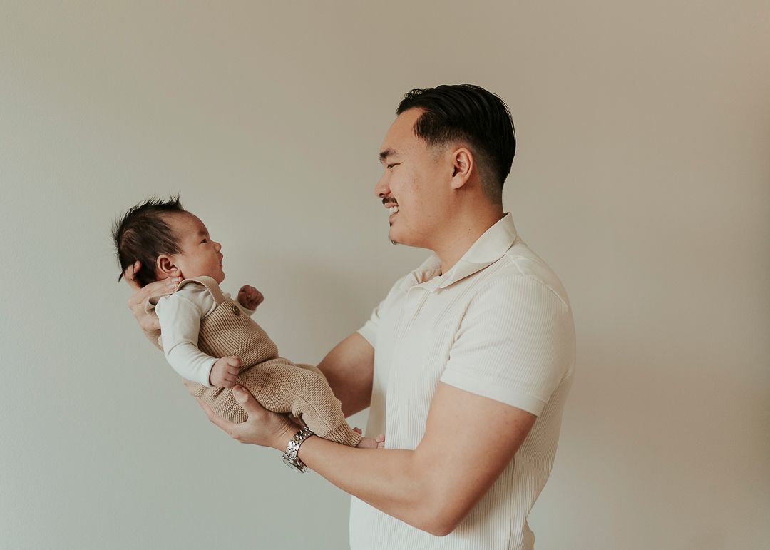 Pappa som står och håller sitt barn, han håller upp barnet framför sig från sidan. De tittar på varandra
